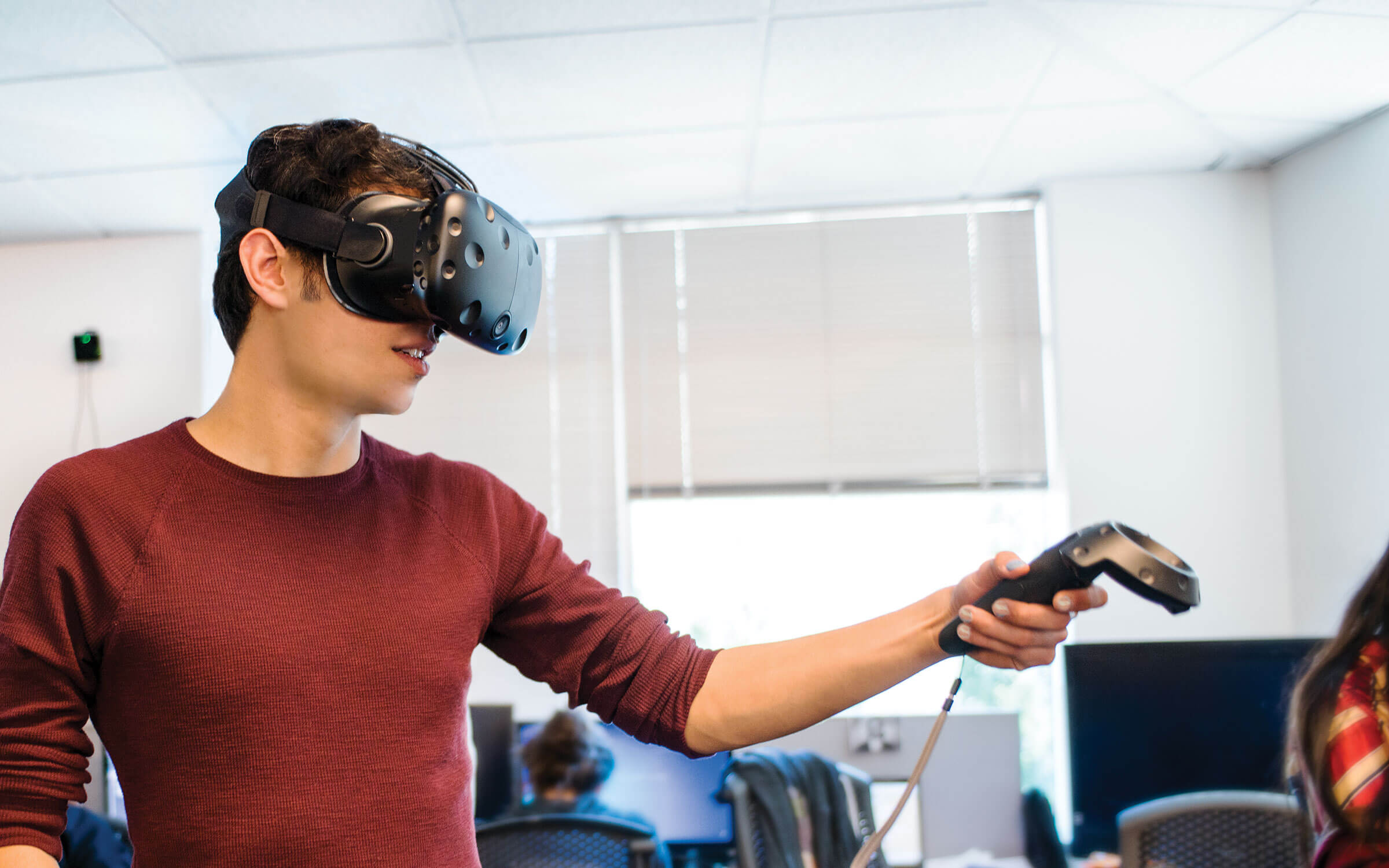 Man wearing virtual reality goggles holds up a game controller in one hand.