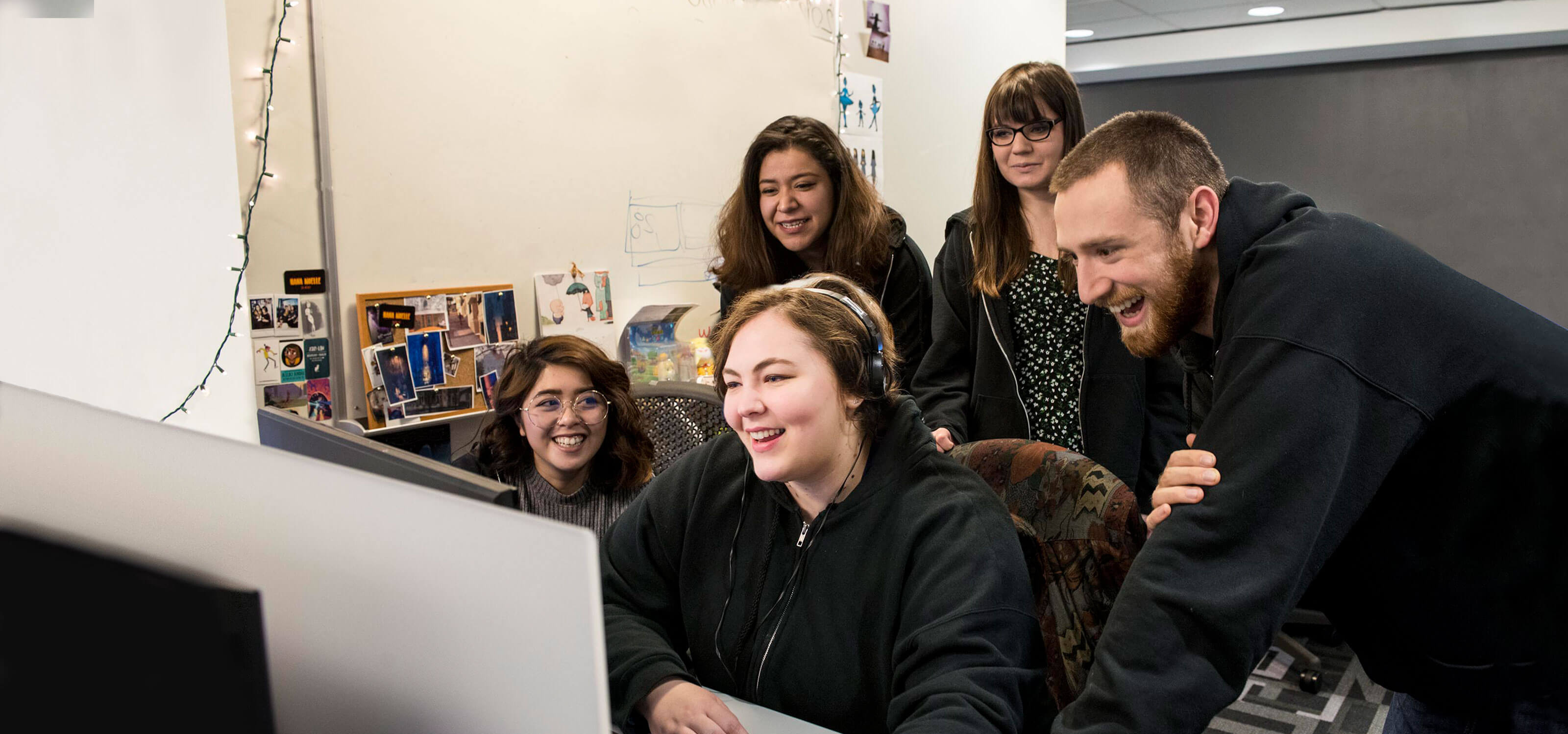 A group of smiling people stand behind a woman sitting at a computer monitor, looking over her shoulder.