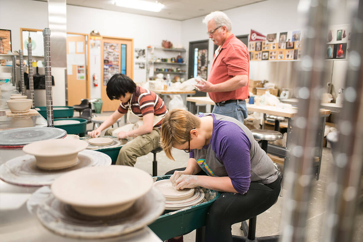 Students work intently at pottery wheels in a ceramics studio as an instructor behind them looks over their shoulders.