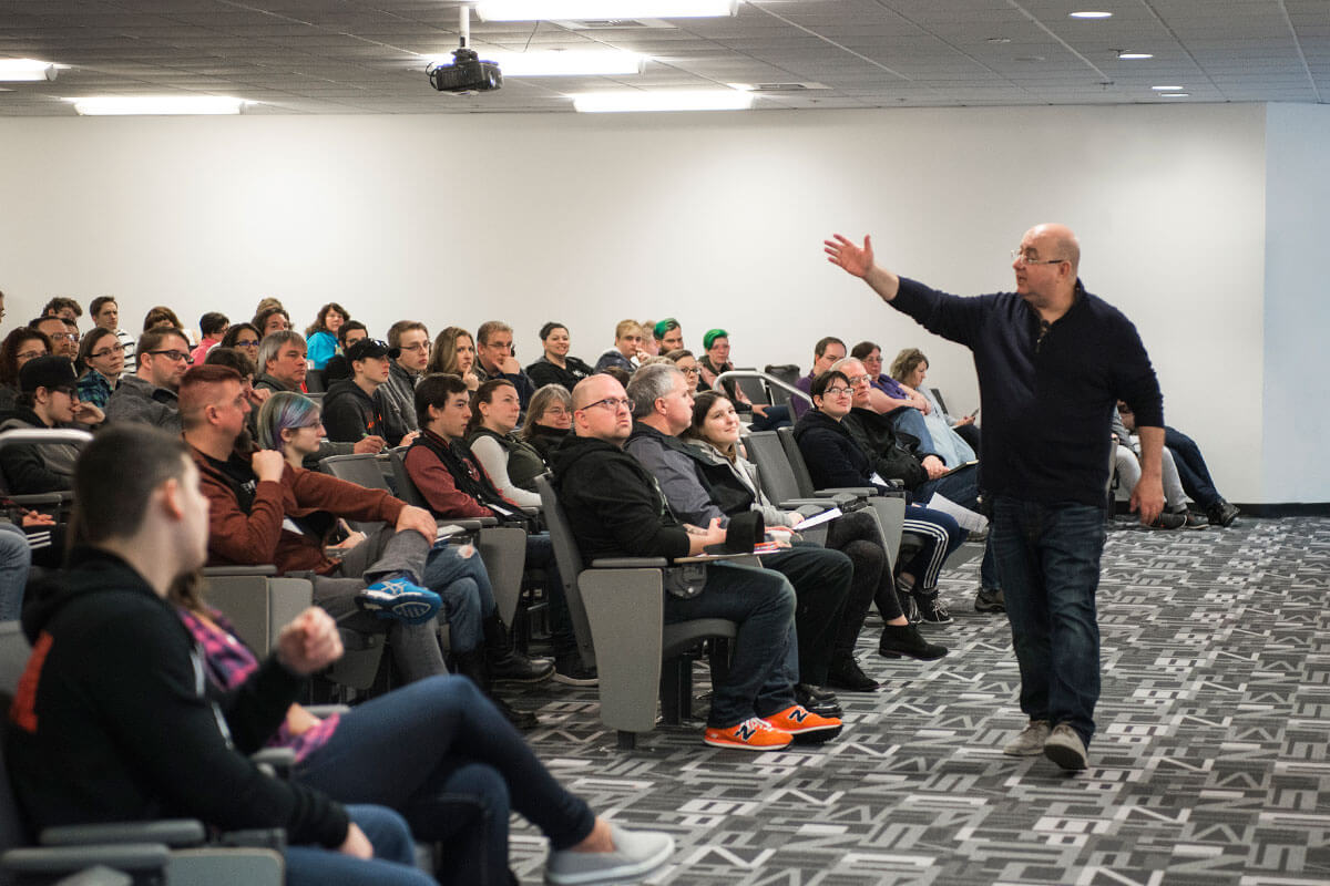 A man in jeans and a black parka animatedly addresses a group of people in a large auditorium-style classroom.