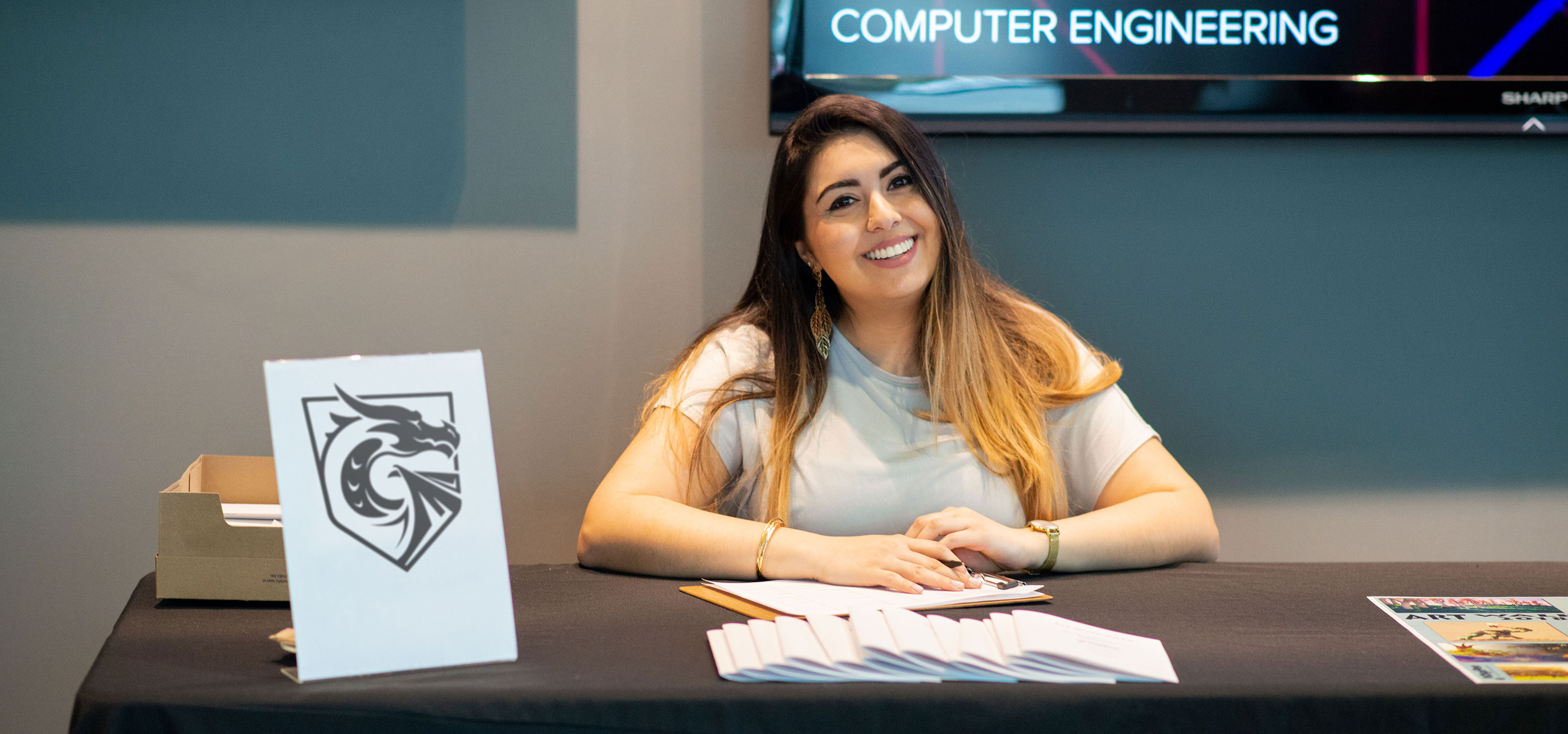 A smiling woman sits at a table welcoming people to an event on DigiPen's campus.