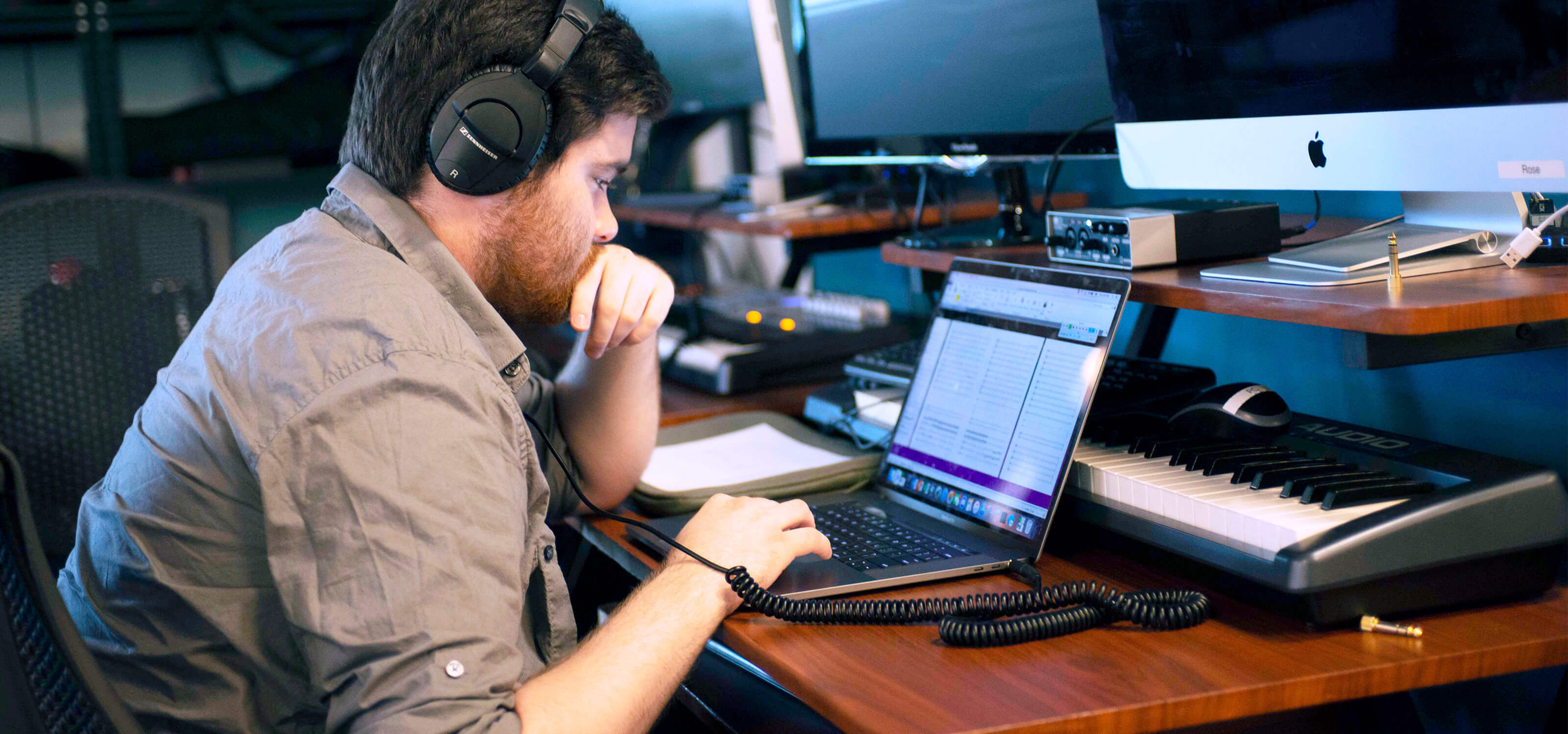 A DigiPen music student wearing headphones works at their laptop in front of a synthesizer.