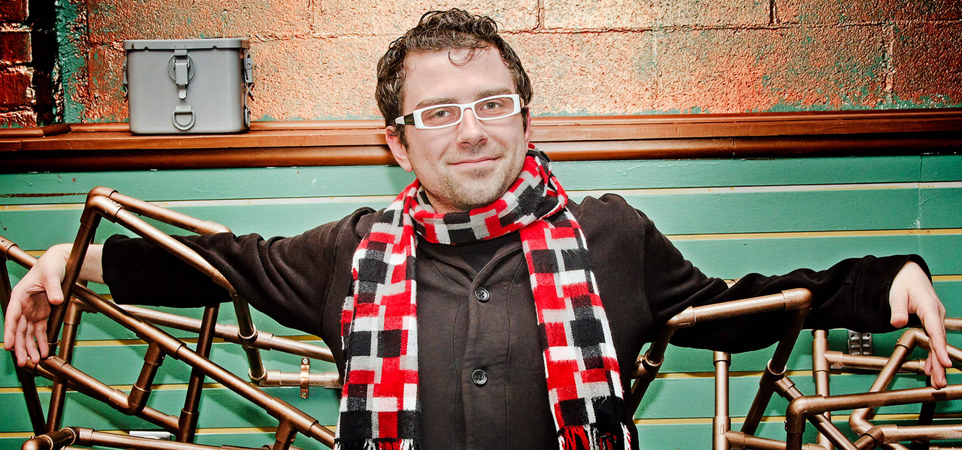 DigiPen alumnus Nate Martin in a plaid scarf, black coat and white-framed glasses with a prop made of bronze-colored pipes