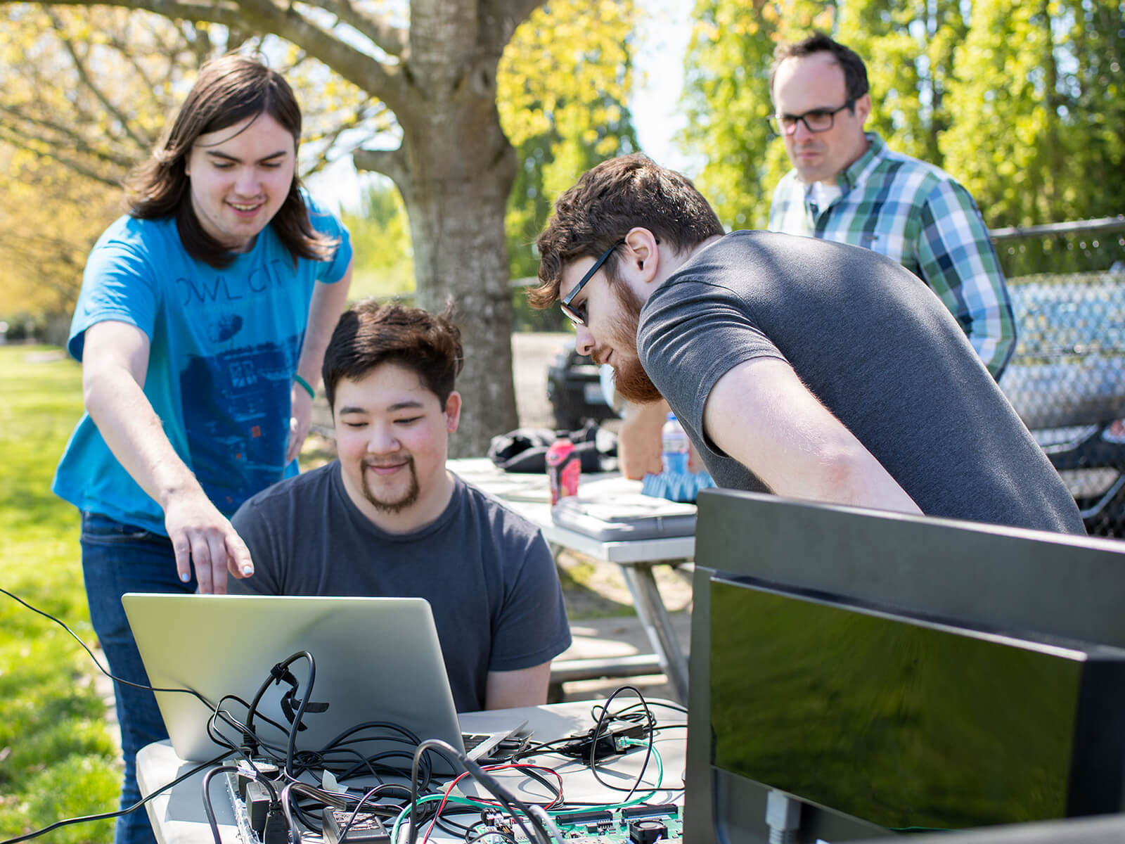 Two students look over the shoulder of another at a laptop computer and radar system set up outdoors in a park.