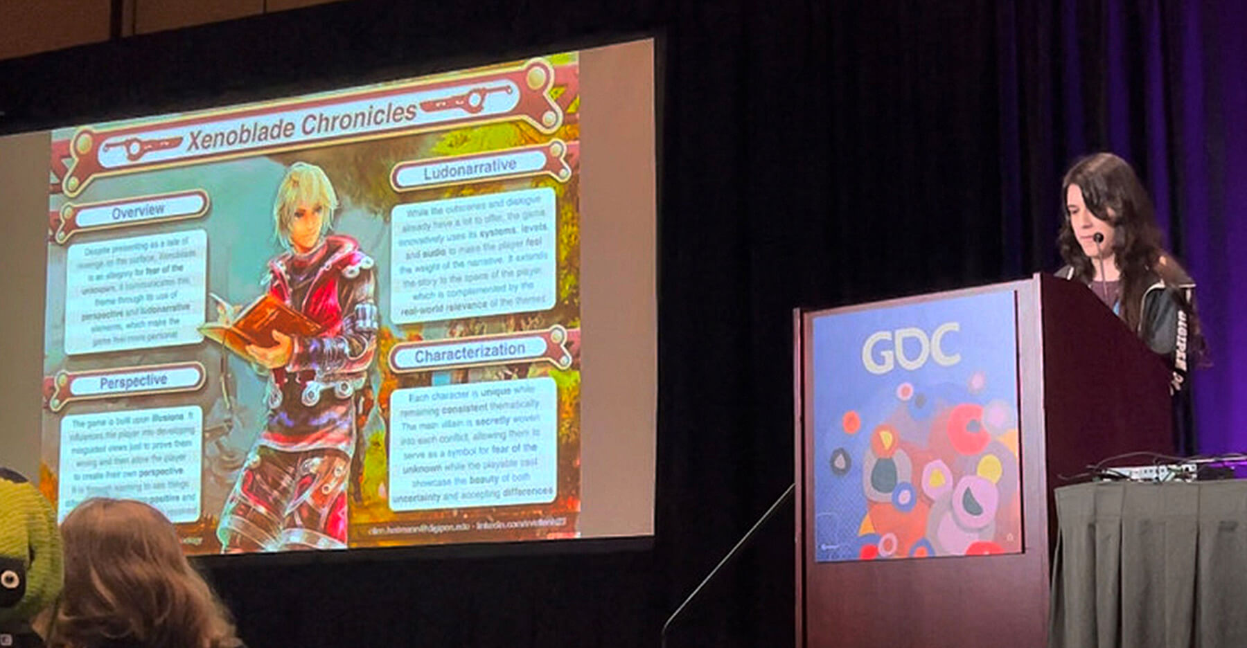 Ellen Heitmann presents at a GDC-branded podium next to a projector screen displaying her Xenoblade Chronicles analysis.