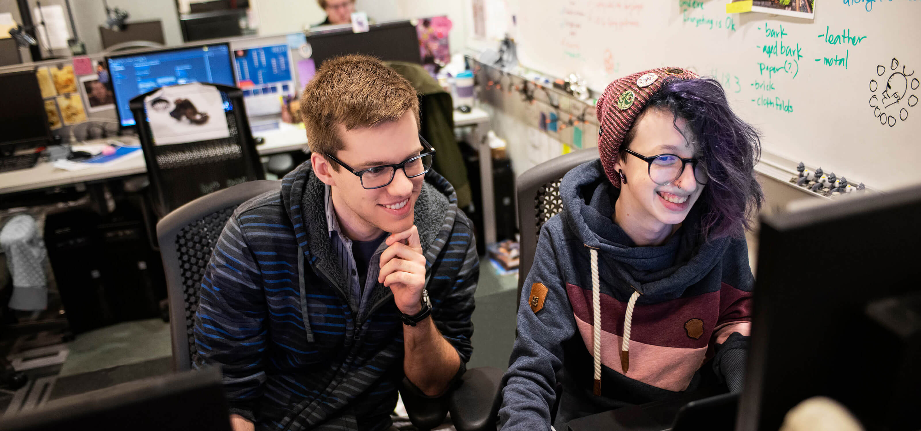 Two students working together in a computer lab looking and smiling at a computer monitor.