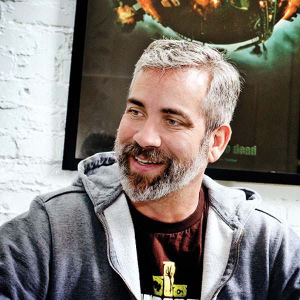 Man with wearing a t-shirt and gray hooded sweater looks to the left, sitting in front of a white wall with a black poster.