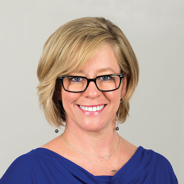 Portrait from the shoulders up of a blond woman wearing black-rimmed eyeglasses against a beige background.