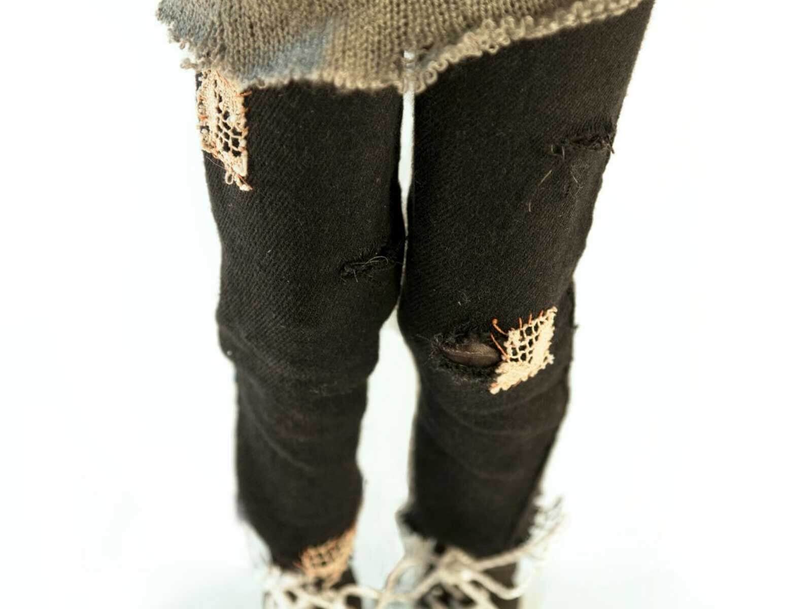 A waist-down shot of a doll’s legs wearing patched up pants.