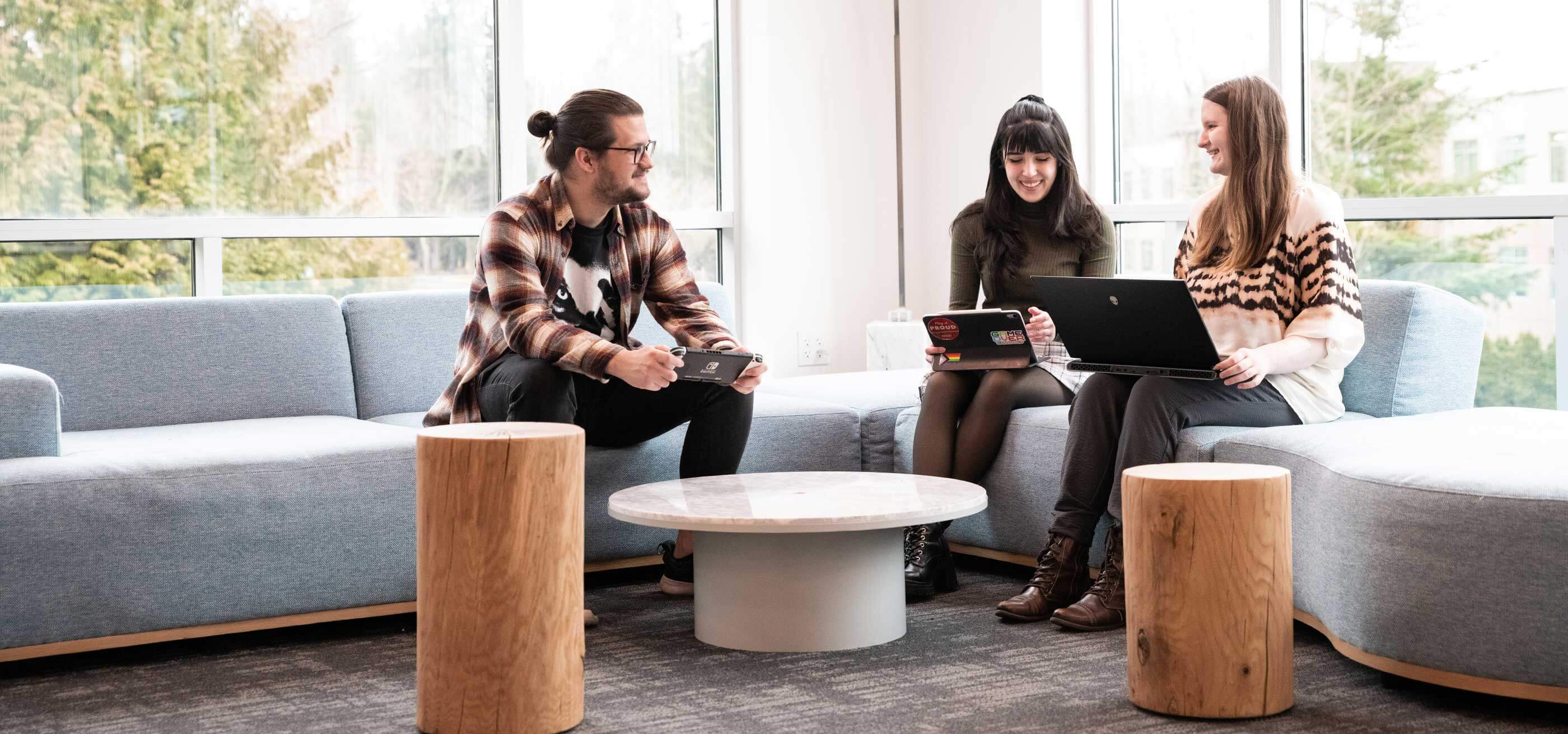 Three students use various electronic devices as they sit and lounge in the library