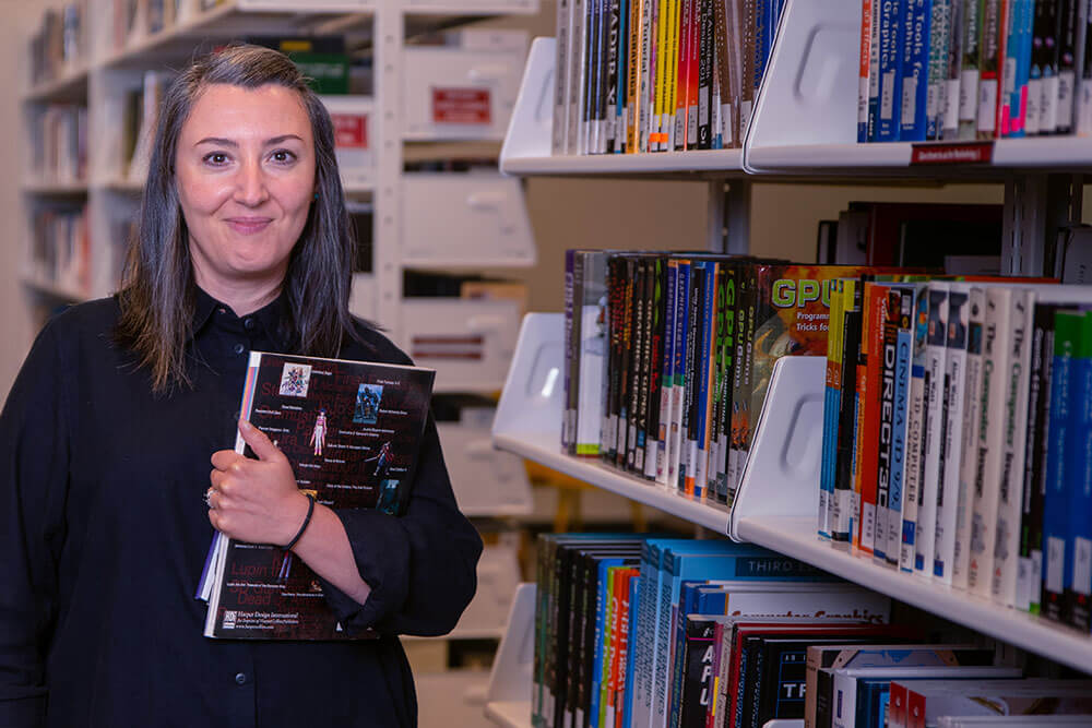 A woman holding books next to a bookshelf.