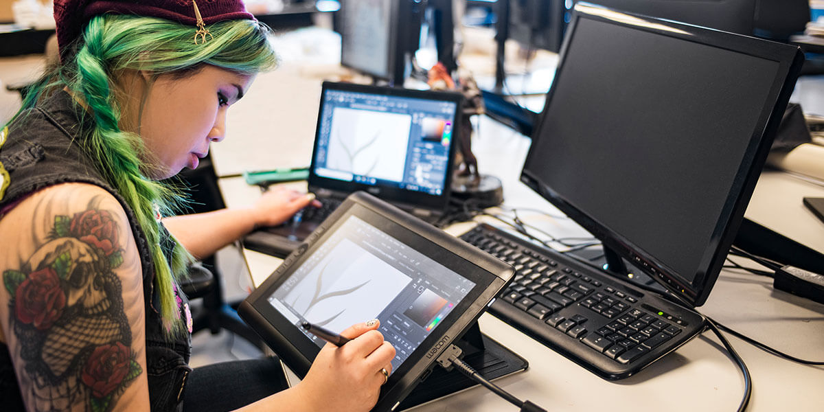 A woman with a skull-and-roses arm tattoo draws on a tablet computer while sitting at a laptop computer and monitor.