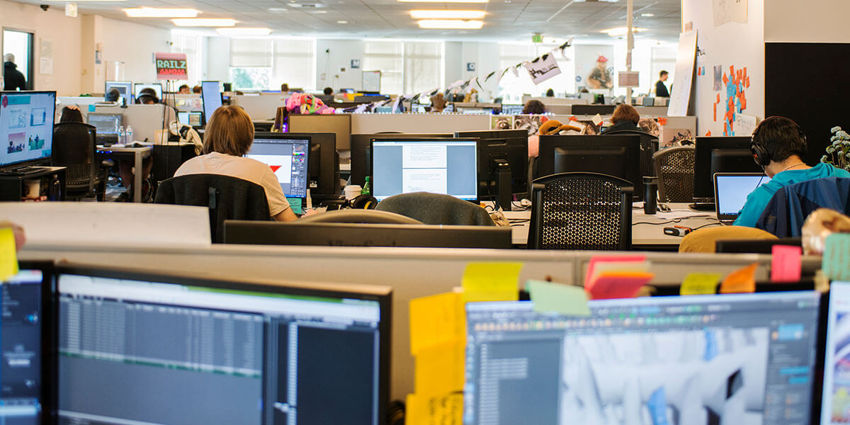 Rows of monitors are seen in a crowded, brightly lit computer lab, some with people sitting at them, working on projects.