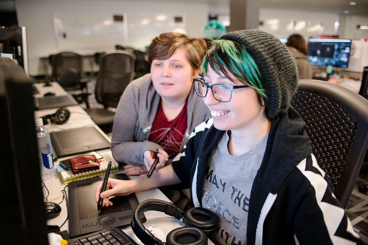 Two students sit in a computer lab looking and smiling at a computer monitor.