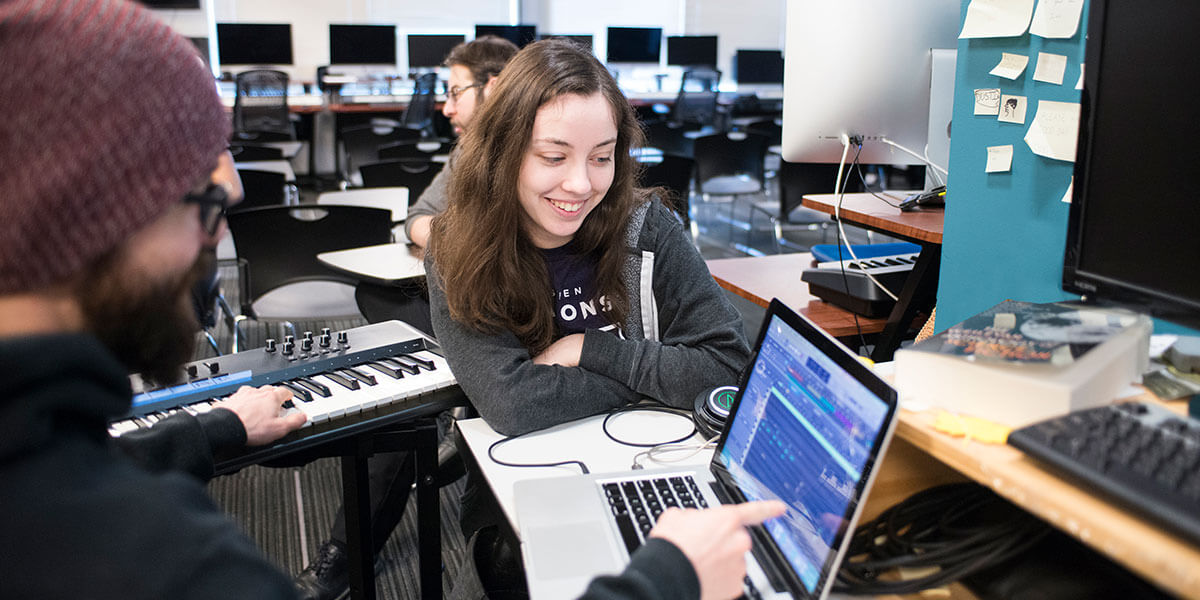 A woman in a gray sweater smiles sitting at a desk next to a man pointing at the screen of a laptop computer.