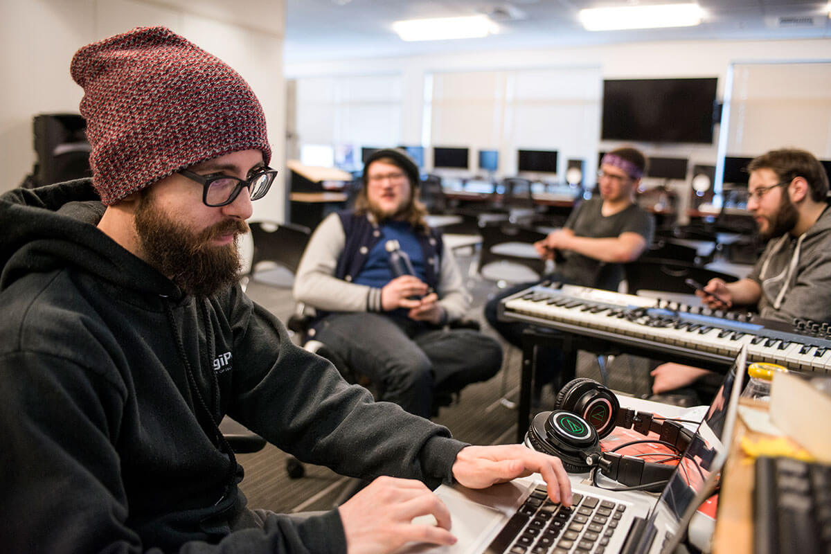 A man in a beanie and black sweater sits at a laptop computer in front of three others next to a standing electronic piano.