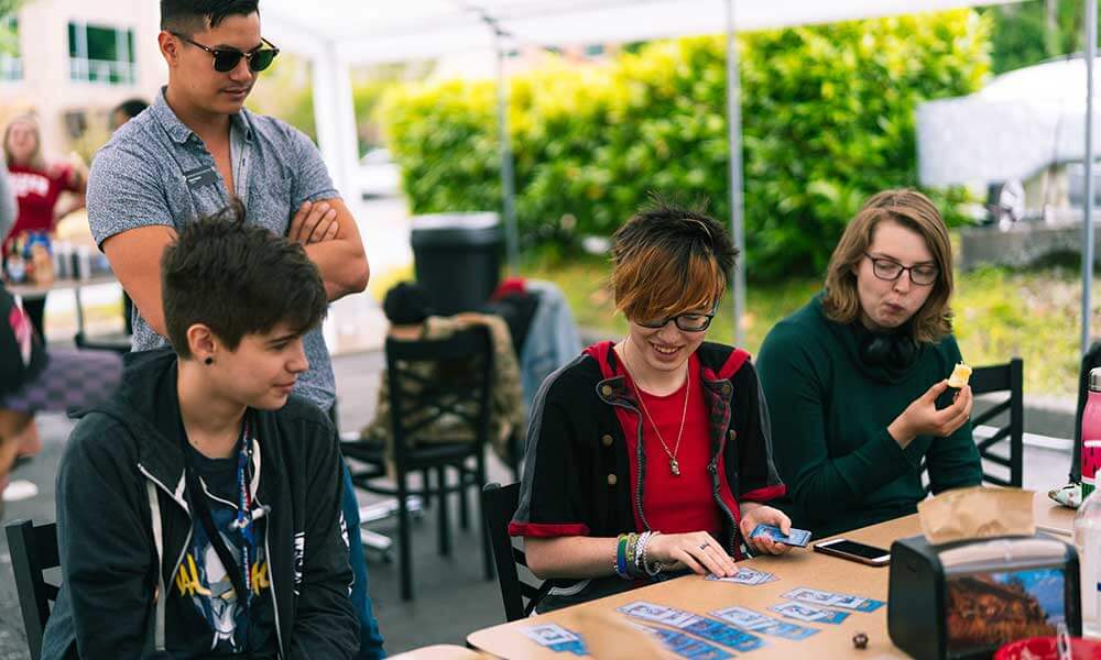 Three students under an outdoor tent watch another student play a card game