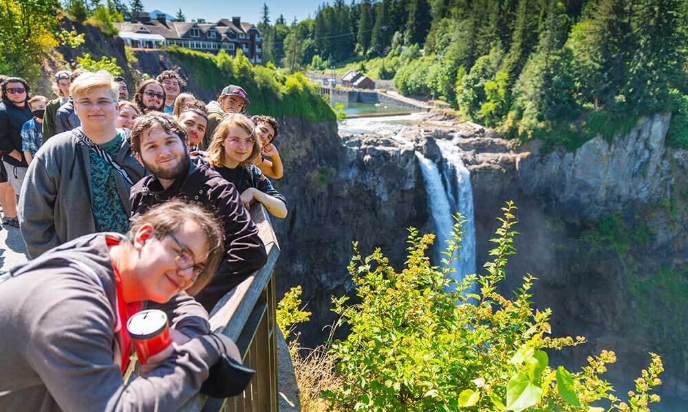 A group of students pose along a handrail with Snoqualmie Falls to their right