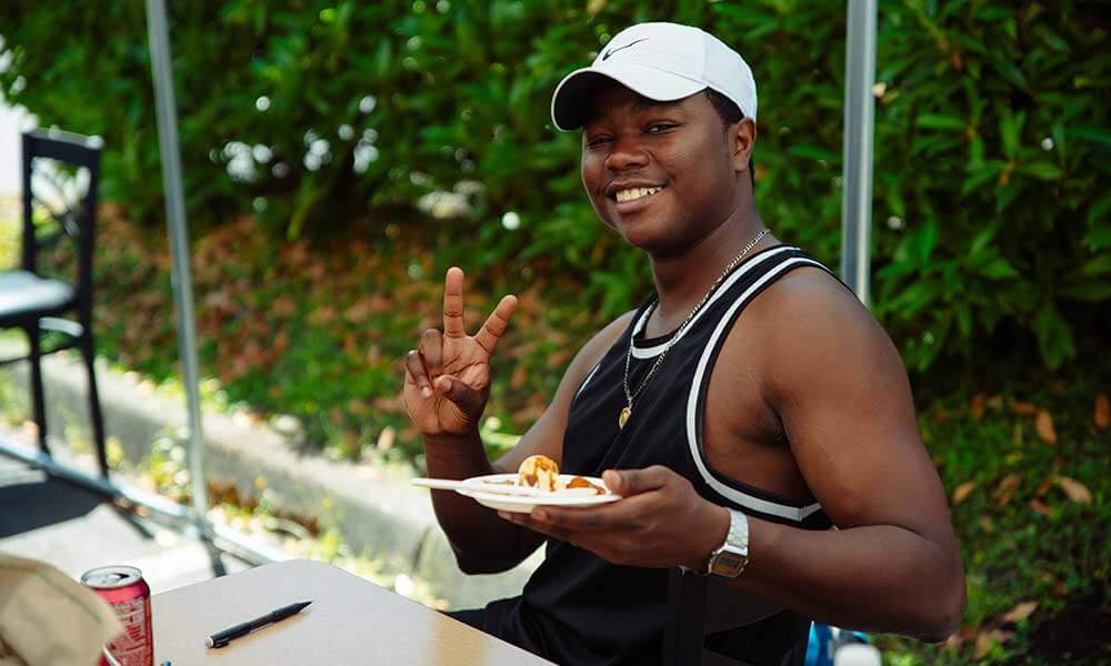 A student in a tank top holding a plate of food waves at the camera