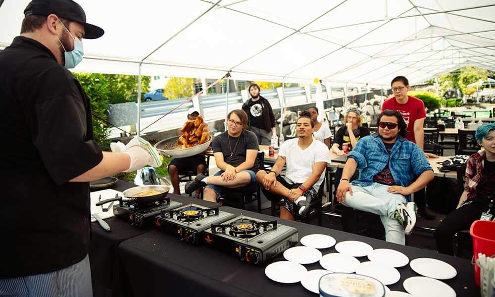 A group of students in an outdoor tent watch a DigiPen chef make their food