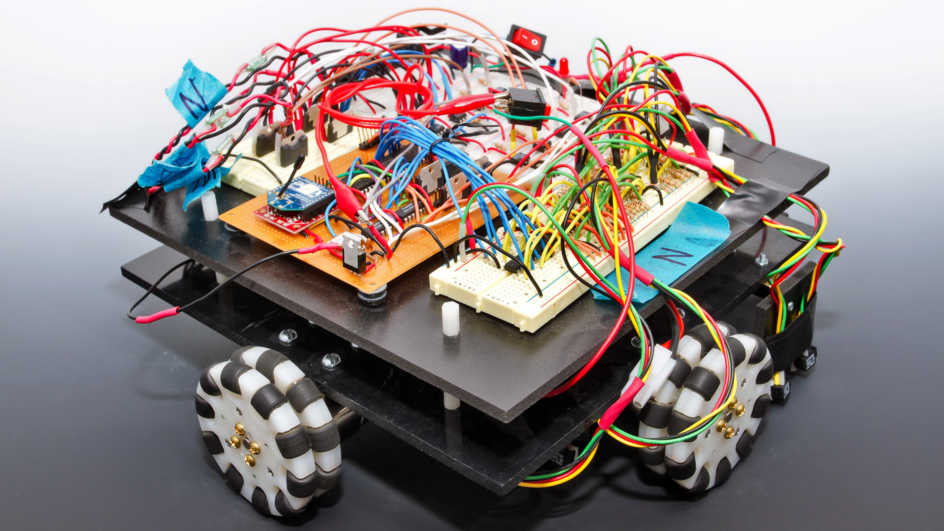 A square robot with exposed circuitry and black and white wheels.