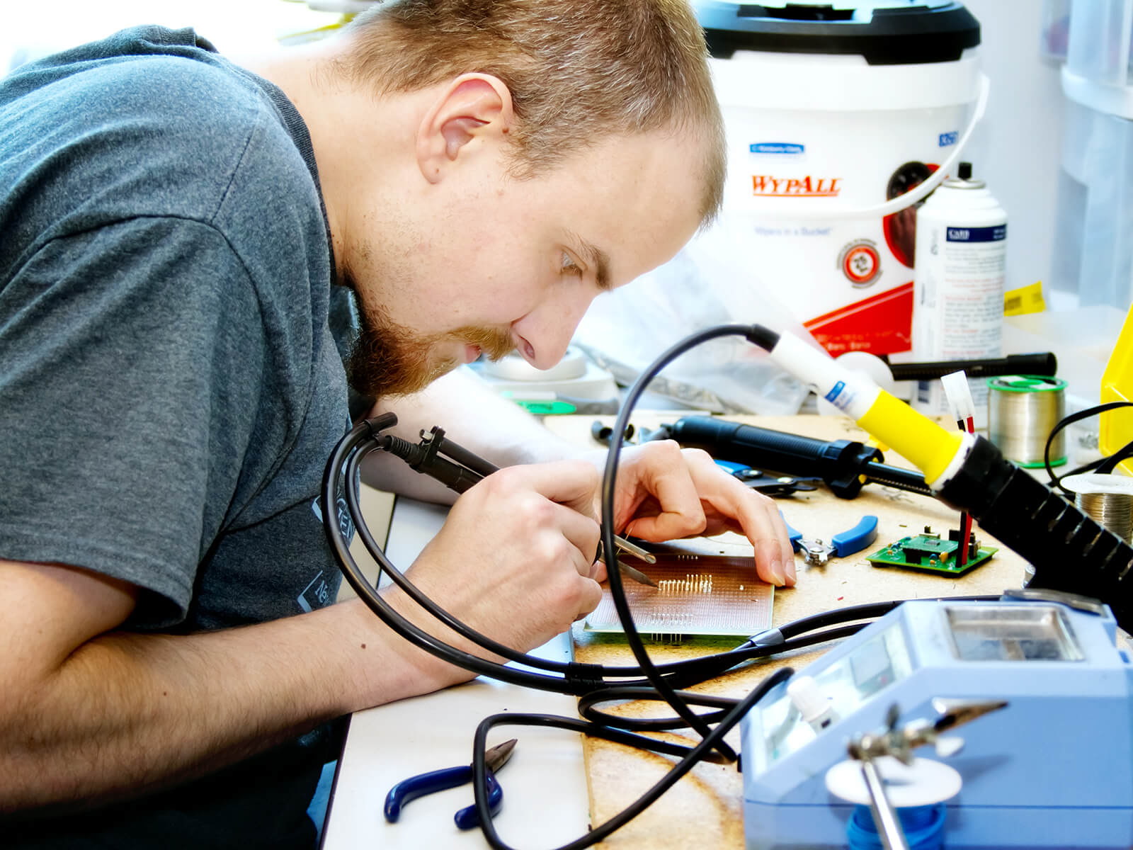 A student solders a circuit board on a desk cluttered with tools.