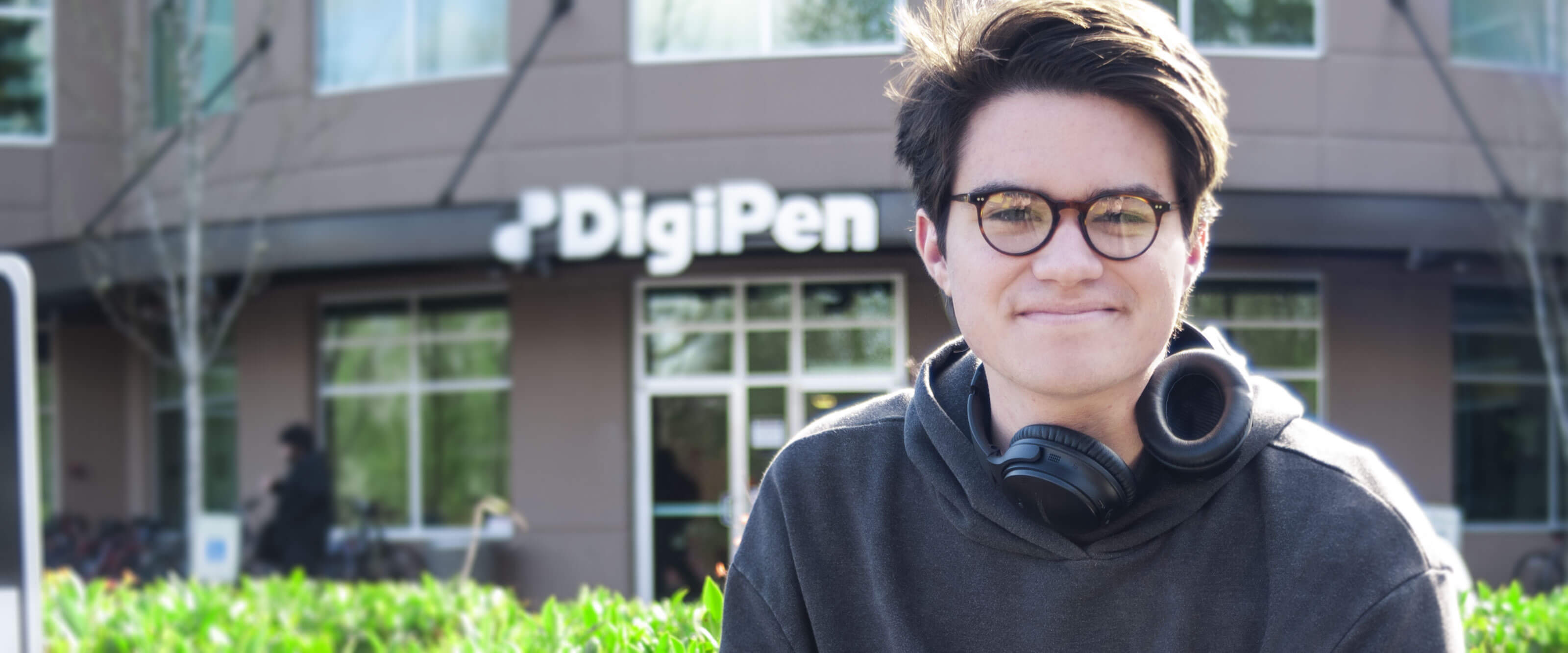 A student wearing headphones smiles on a sunny day in front of the DigiPen building.