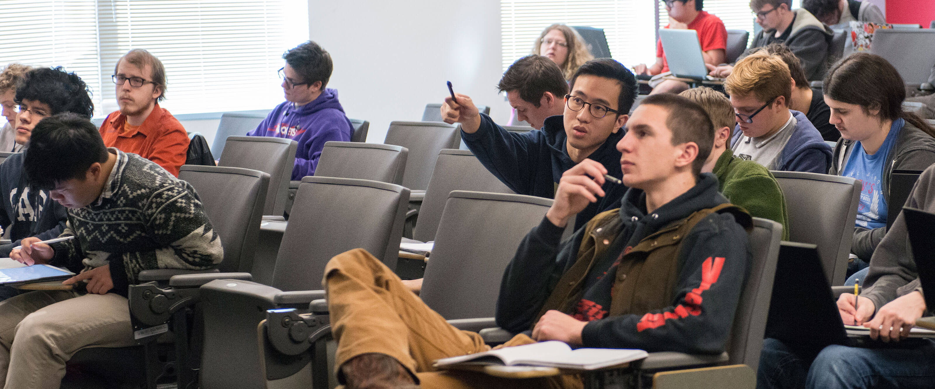 Students sit in a brightly lit auditorium-style classroom taking notes or looking toward the front.