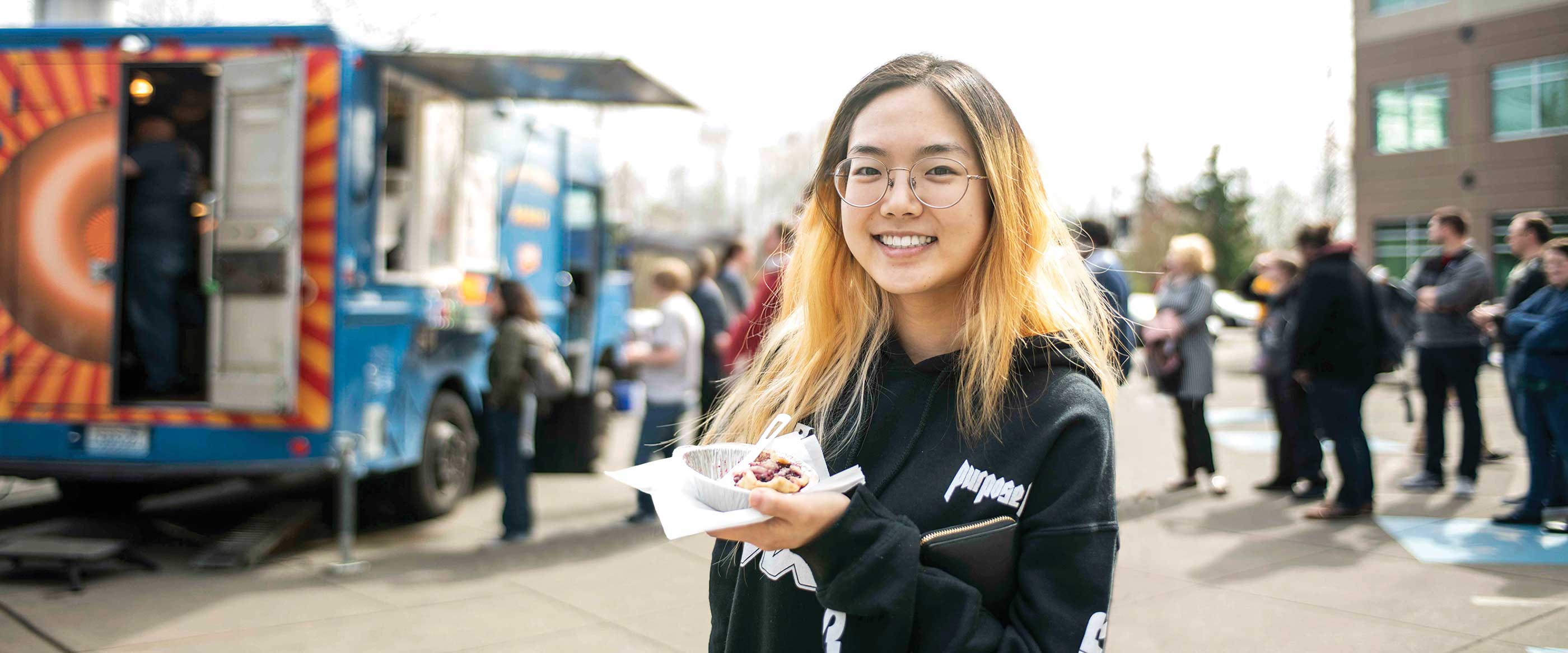 A DigiPen student smiles while eating pie in front of a food truck on campus.