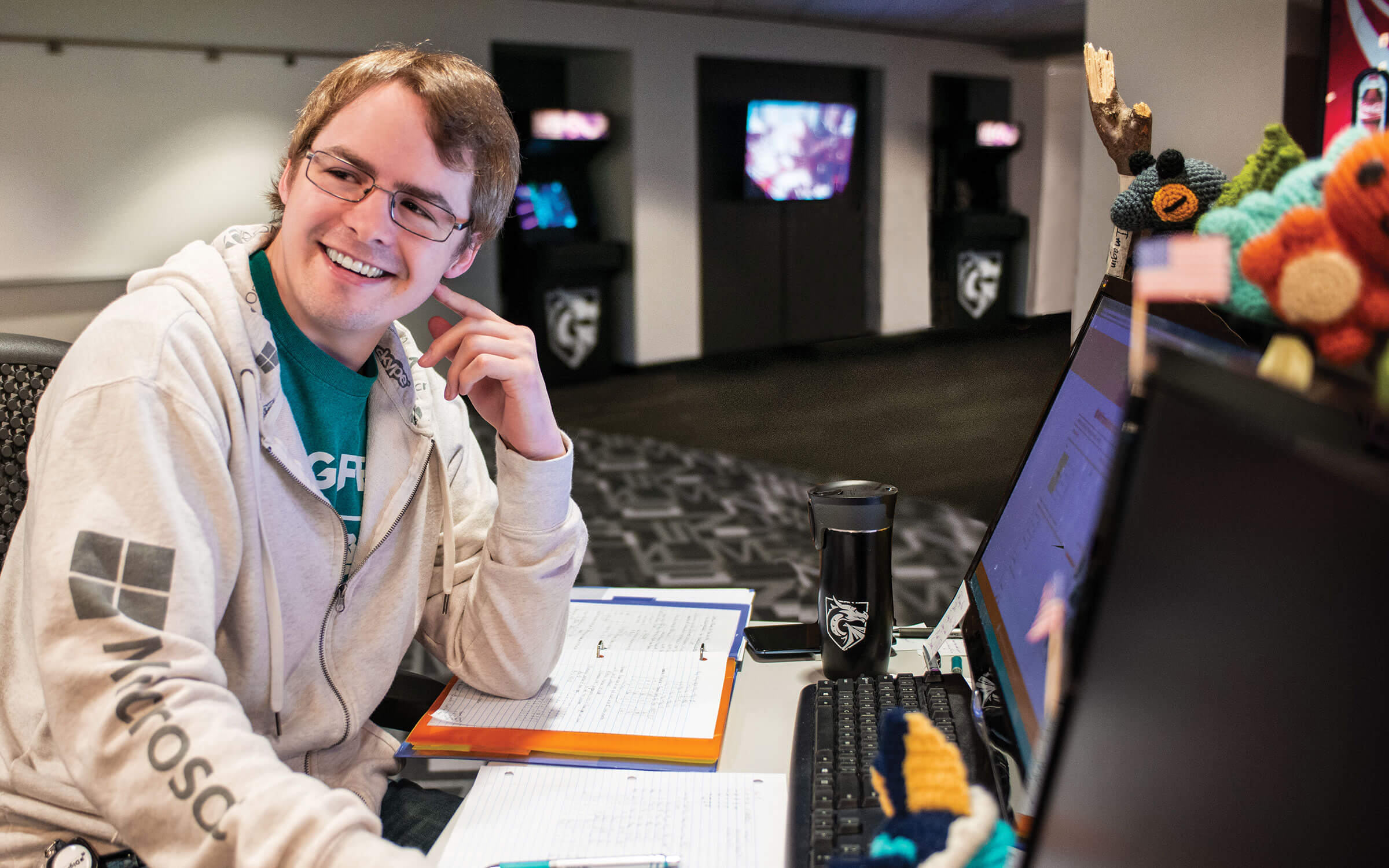 A student at his computer smiles with a DigiPen coffee mug and class notes in front of him.
