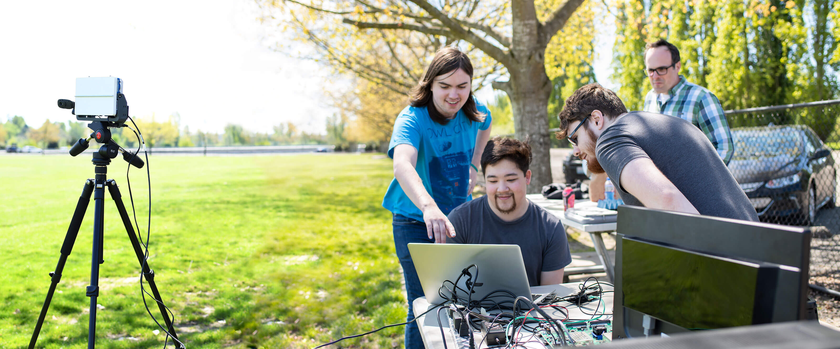 Two students look over the shoulder of another at a laptop computer and radar system set up outdoors in a park.