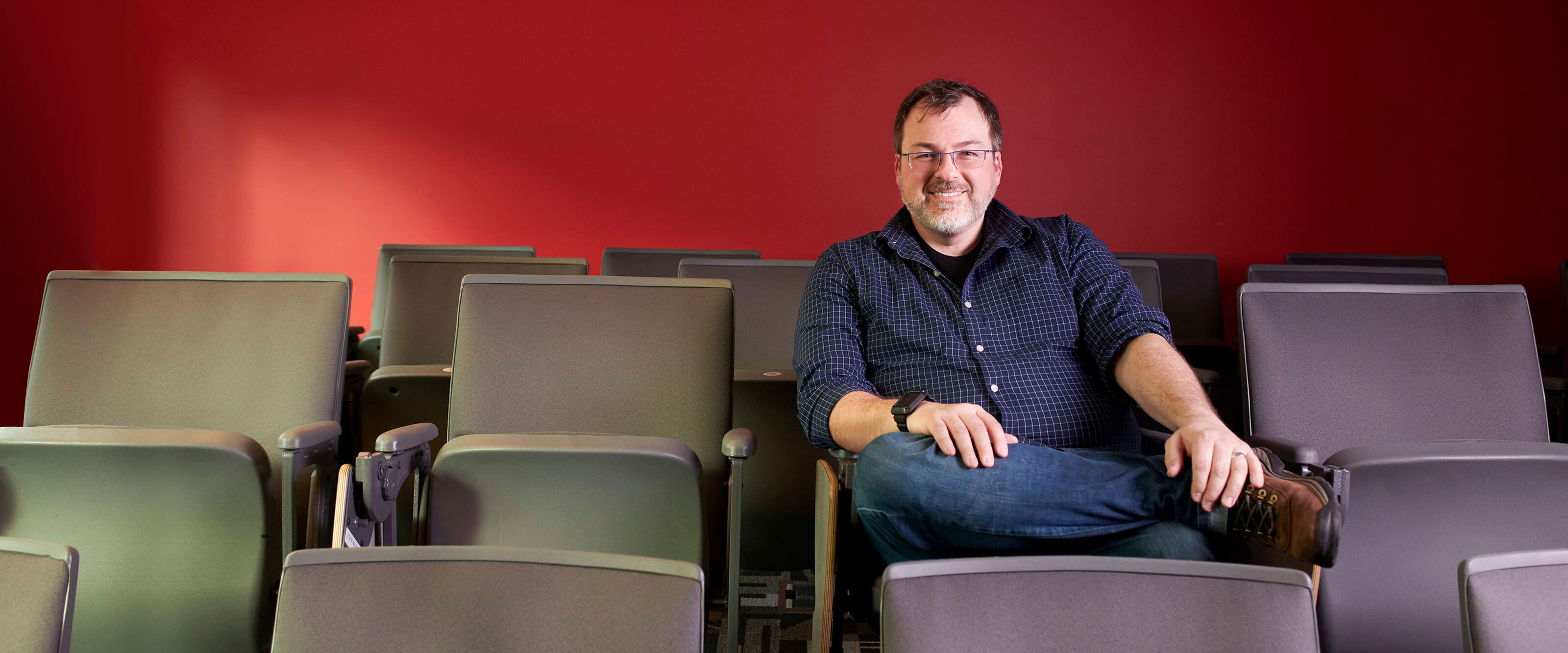 Seen from the front, a man in a blue-patterned shirt sits in a lecture hall in front of a red wall.