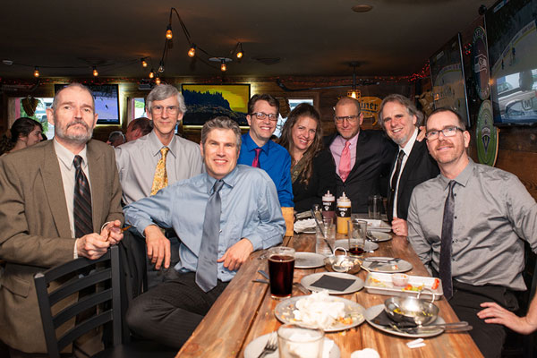 A group of people in formal dress pose for a photo around a table of food and drink in a restaurant.