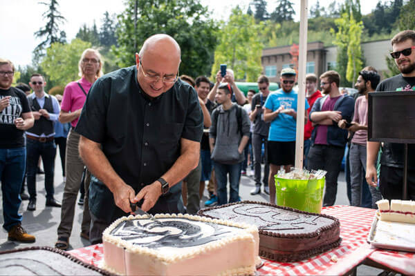 DigiPen president Claude Comair underneath a tent outside cuts a cake depicting the DigiPen Dragon logo while a crowd watches from behind.