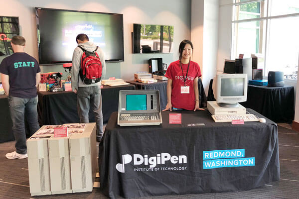 A woman in a red DigiPen t-shirt stands at a convention table flanked by obsolete computer systems and gaming consoles.