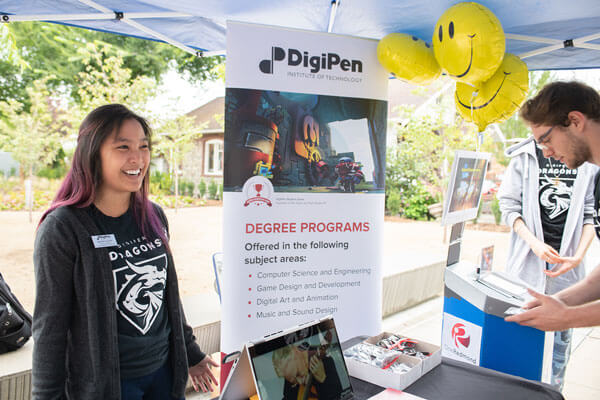 An employee in a black DigiPen Dragons t-shirt stands underneath a tent next to a DigiPen degree program banner.