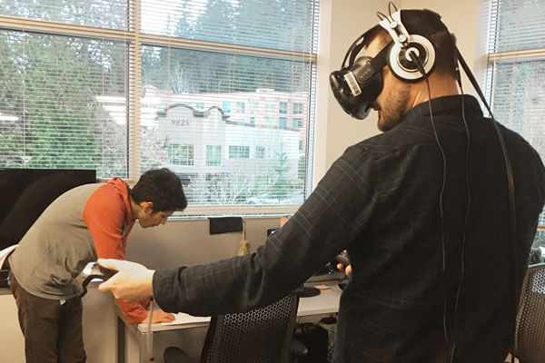 A man wearing a VR headset stands in a classroom gesturing to the left while holding two game controllers.