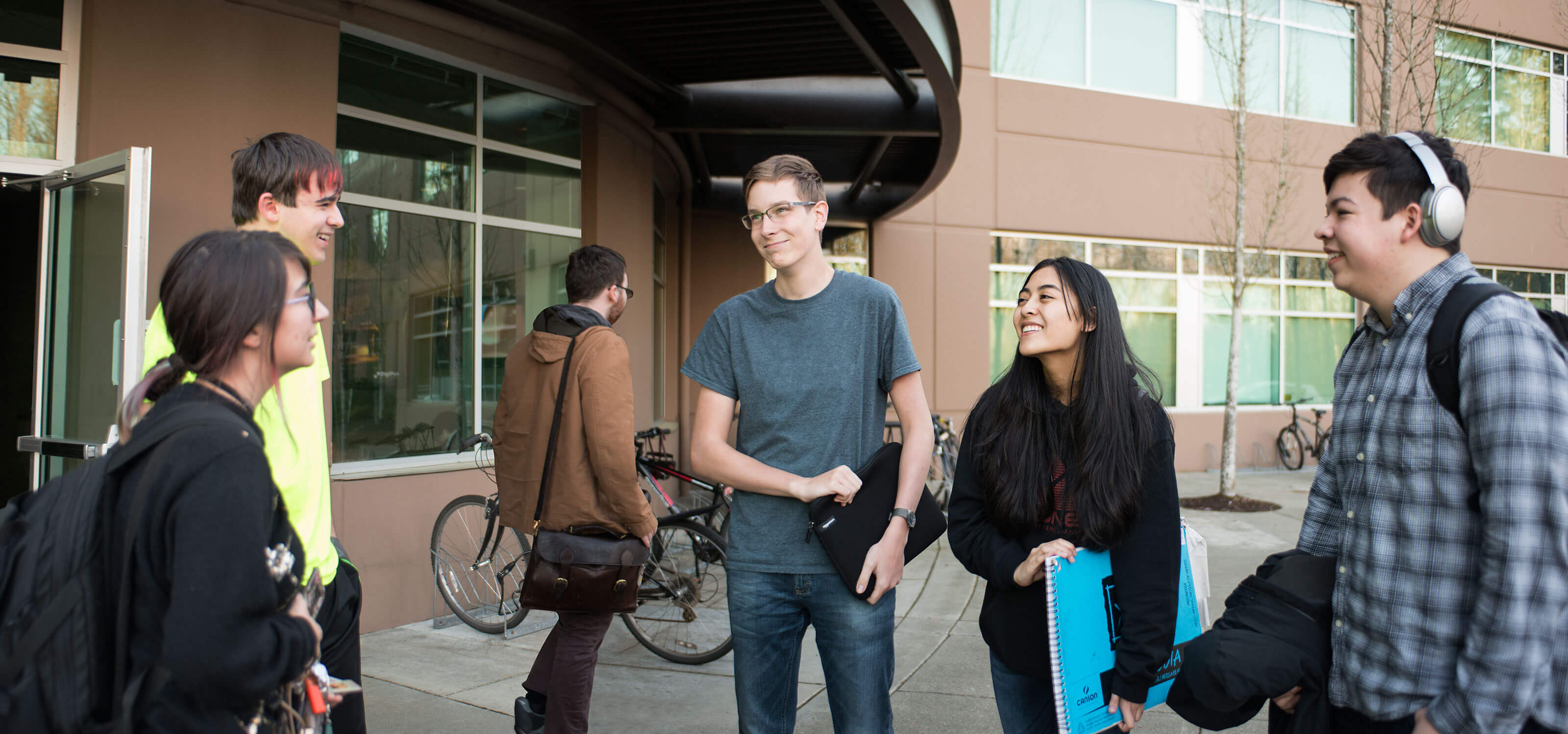 A group of students stand smiling at each other in front of the first-floor lobby of a brown building.