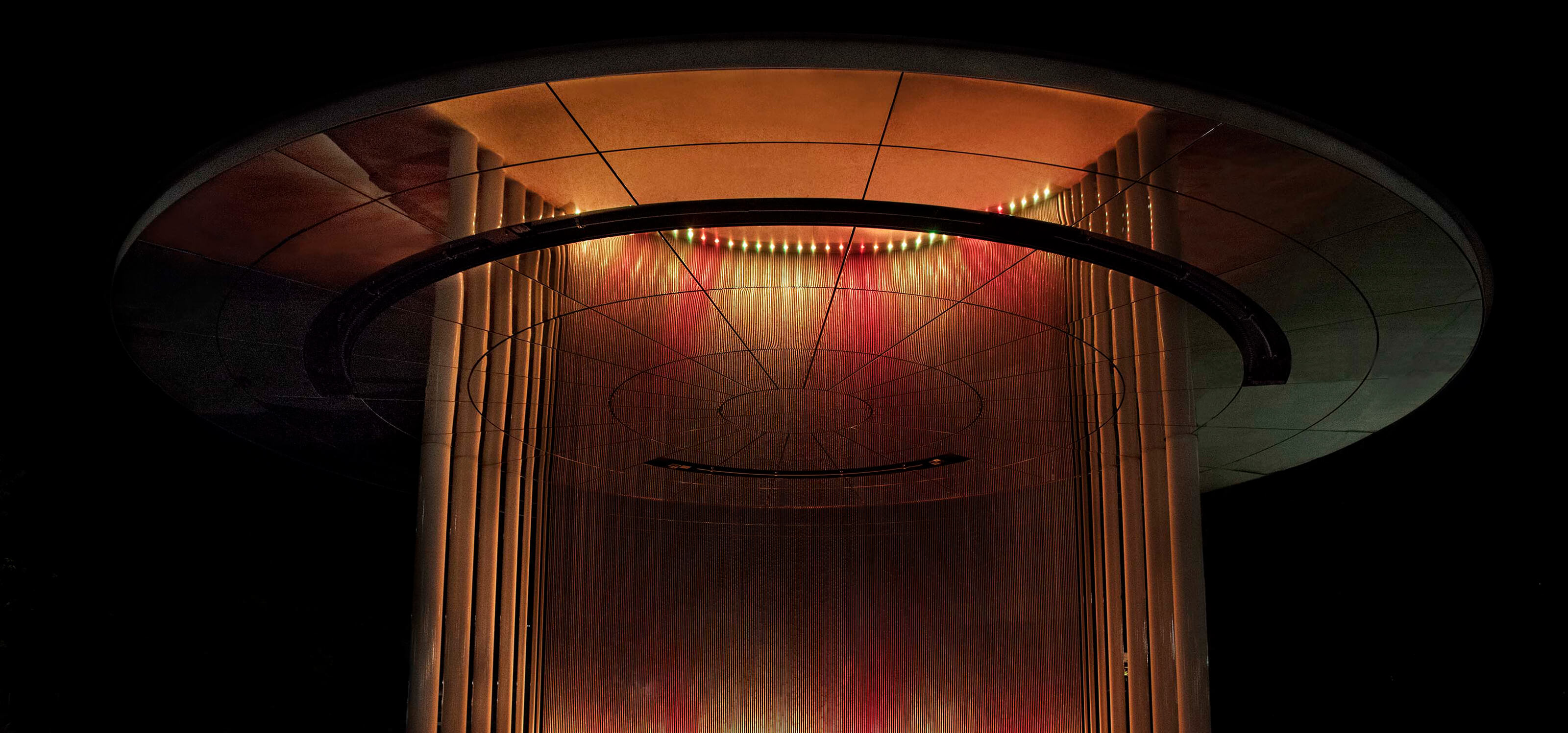 A circular structure with 380 etched metal rods lit in shades of red and yellow at night