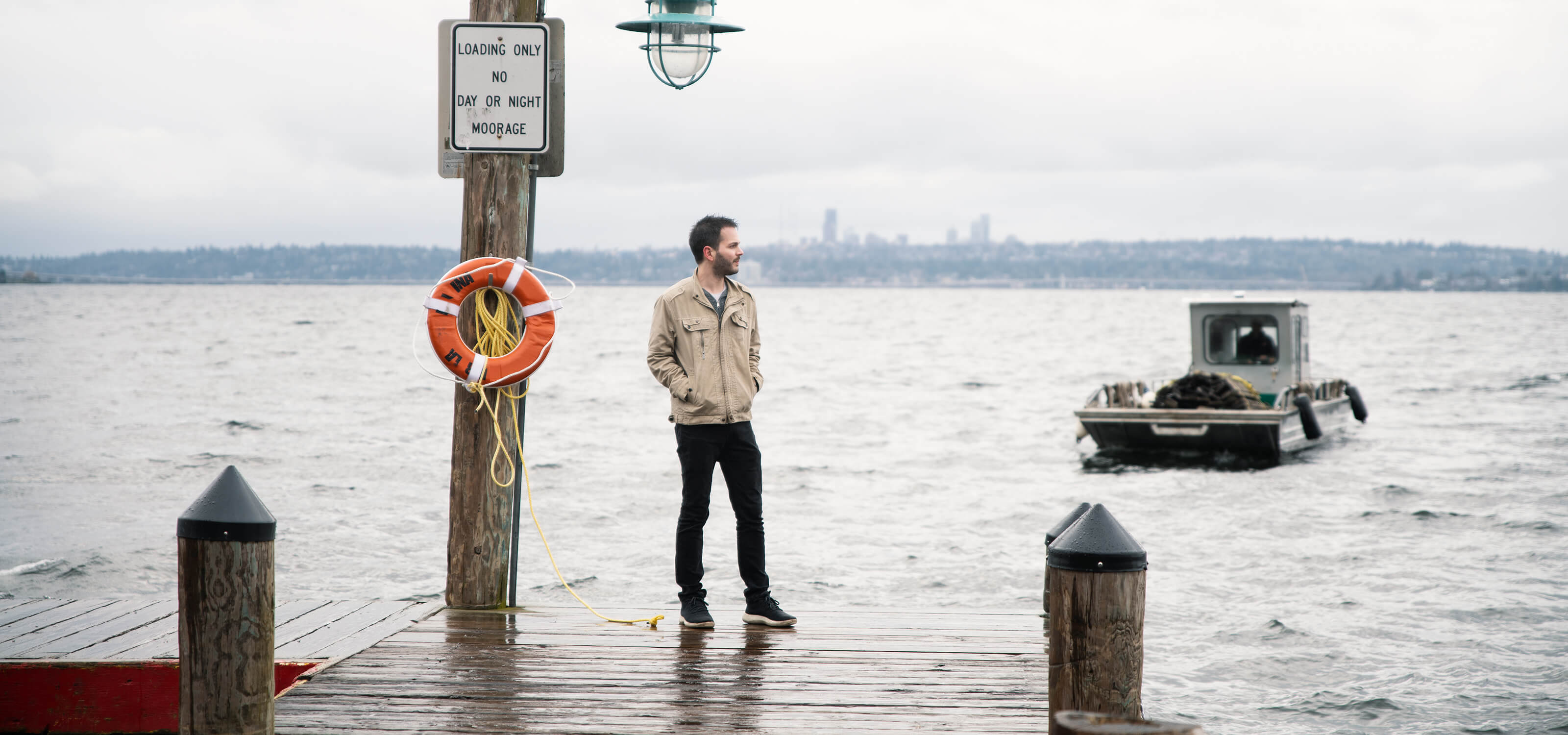 David Evans stands on the end of a dock with small boat and Seattle skyline in background.