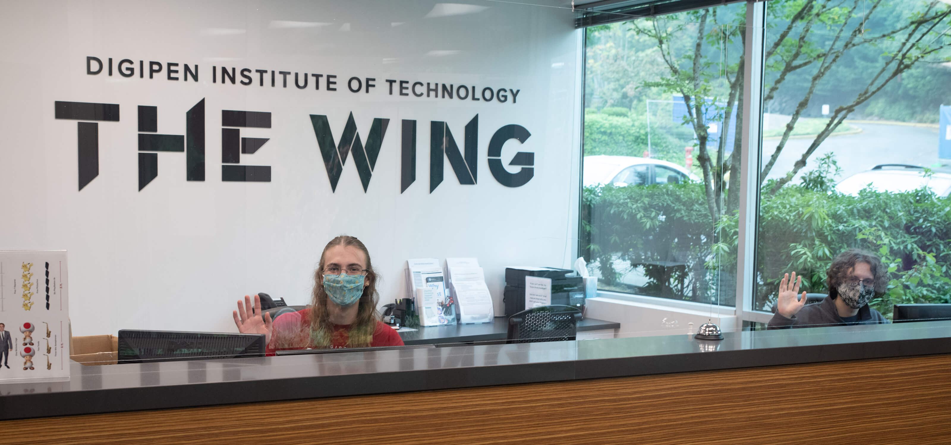 Two masked students wave at the front desk of DigiPen’s new campus expansion, The Wing. 