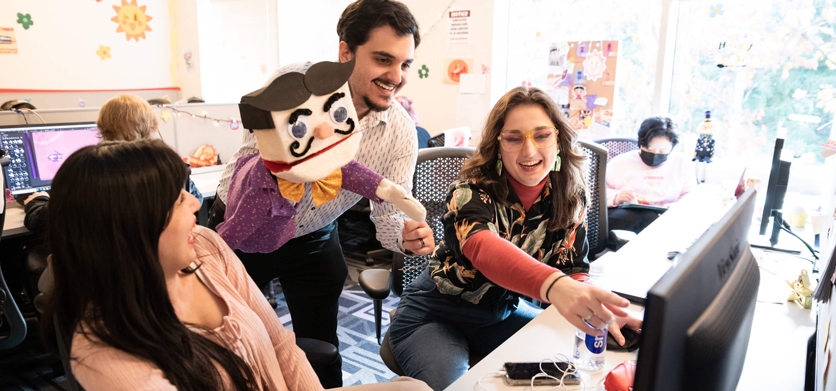 Three DigiPen students, one holding a puppet, smile as they look at a computer in the campus production lab.