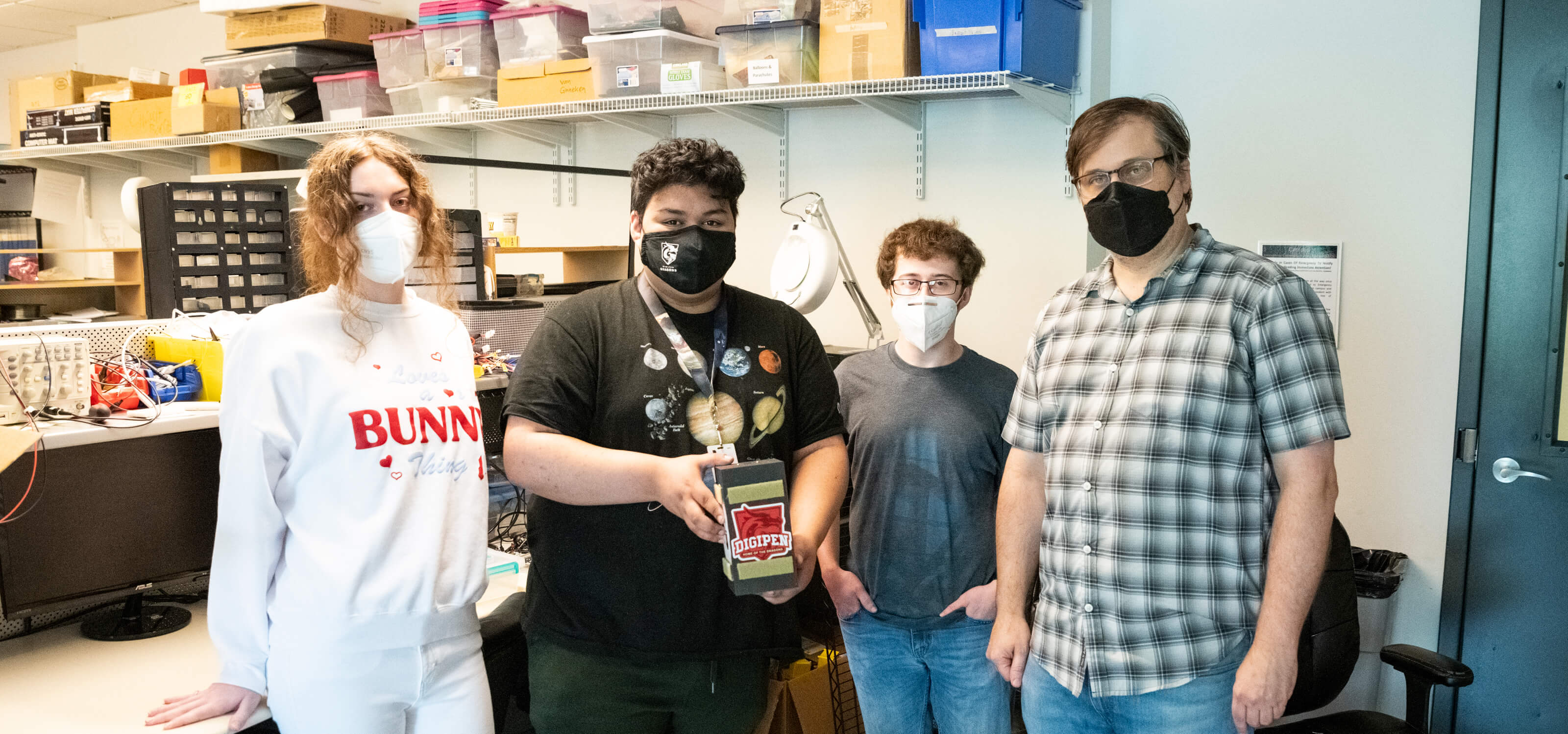 Three students and one professor stand together with student in center holding small payload box.