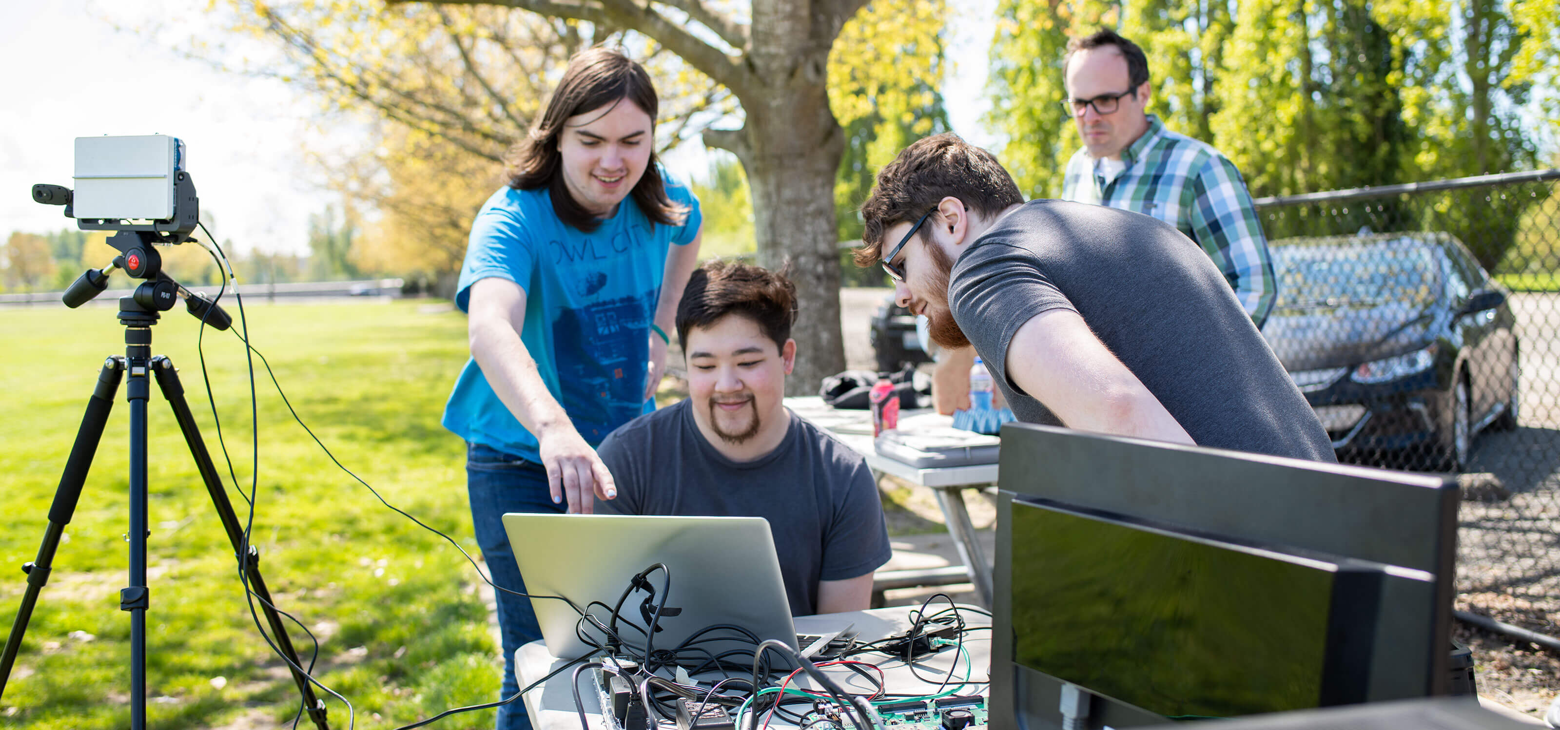 Two students look over the shoulder of another at a laptop computer and radar system set up outdoors in a park.