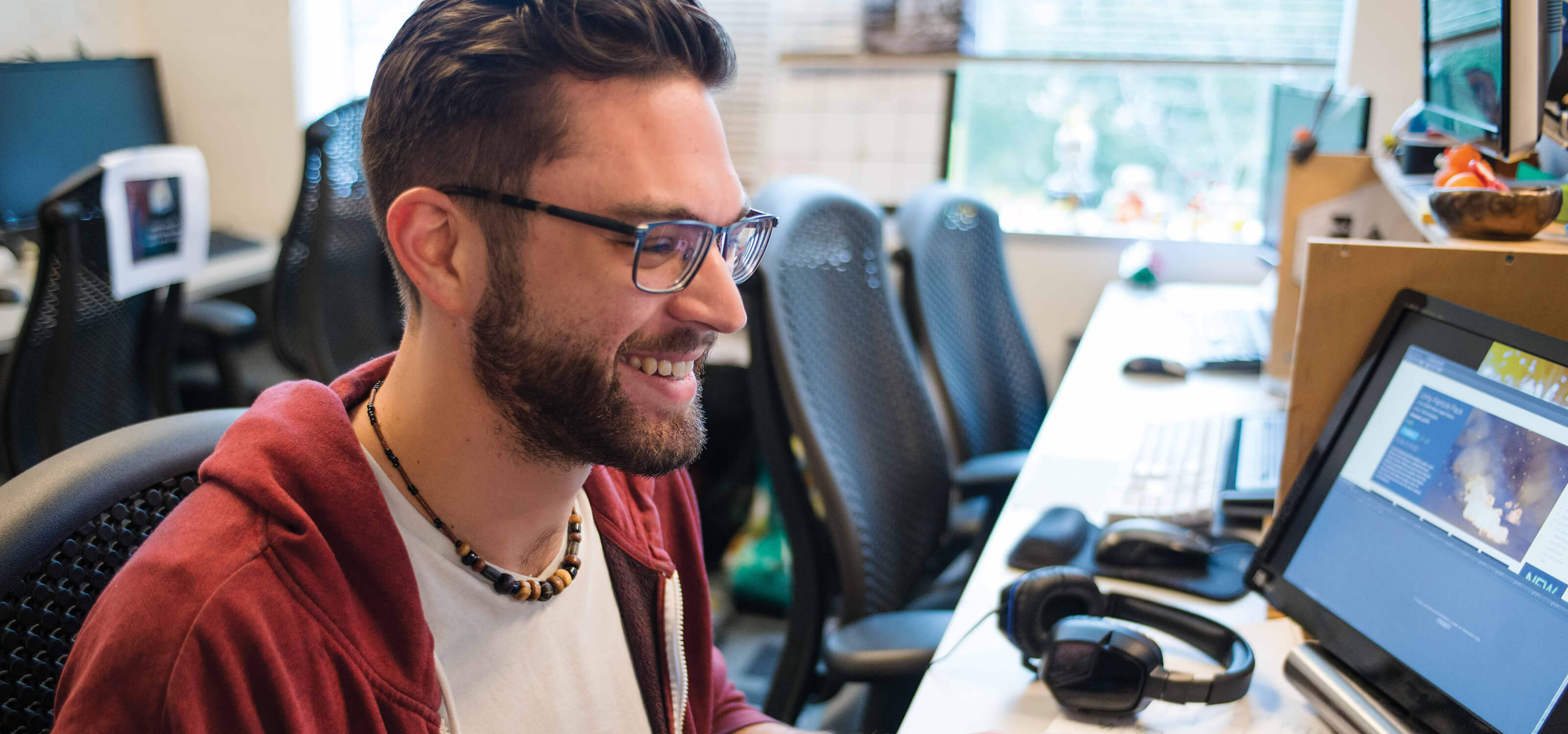 A student wearing glasses sits in a computer lab smiling at his computer screen.