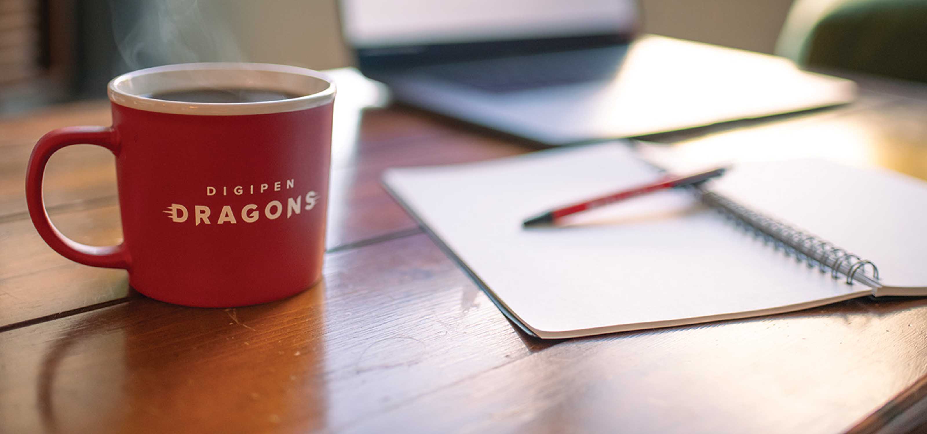 A filled DigiPen branded coffee cup sits next to an open notebook and laptop