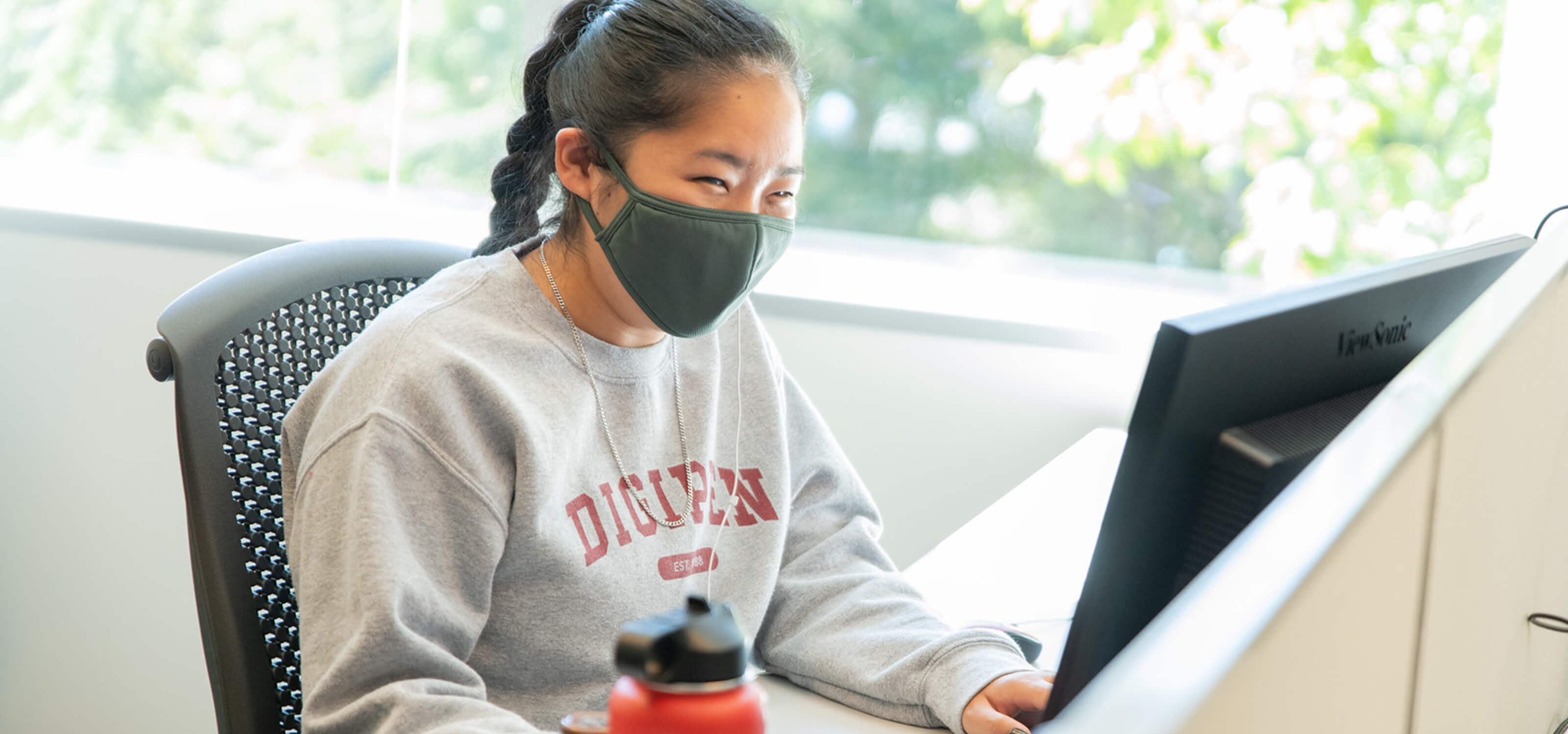 A student wearing a facemask and DigiPen sweatshirt sits in front of a computer screen.