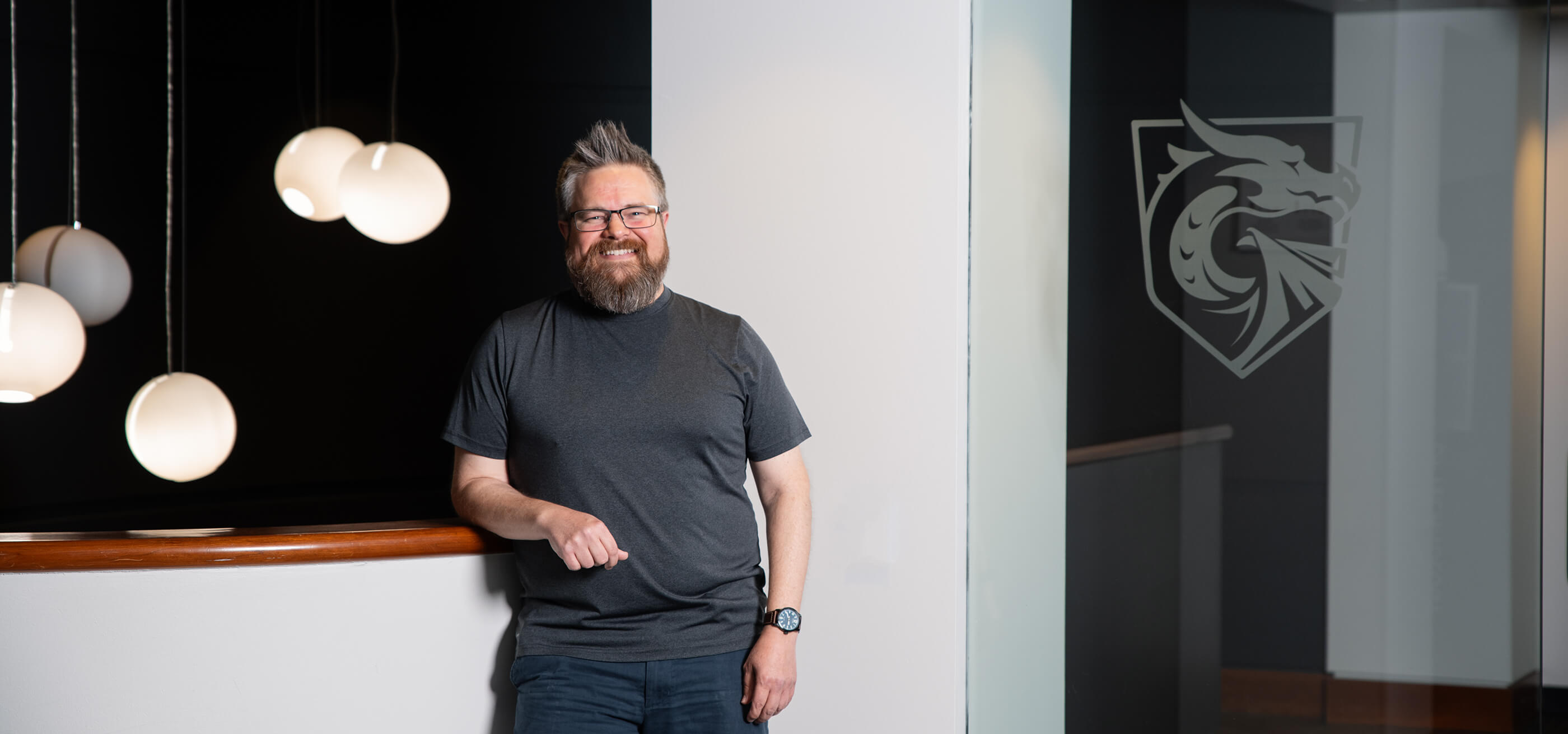 Chris Poplin smiles on campus standing next to a DigiPen Dragons logo decal on glass.