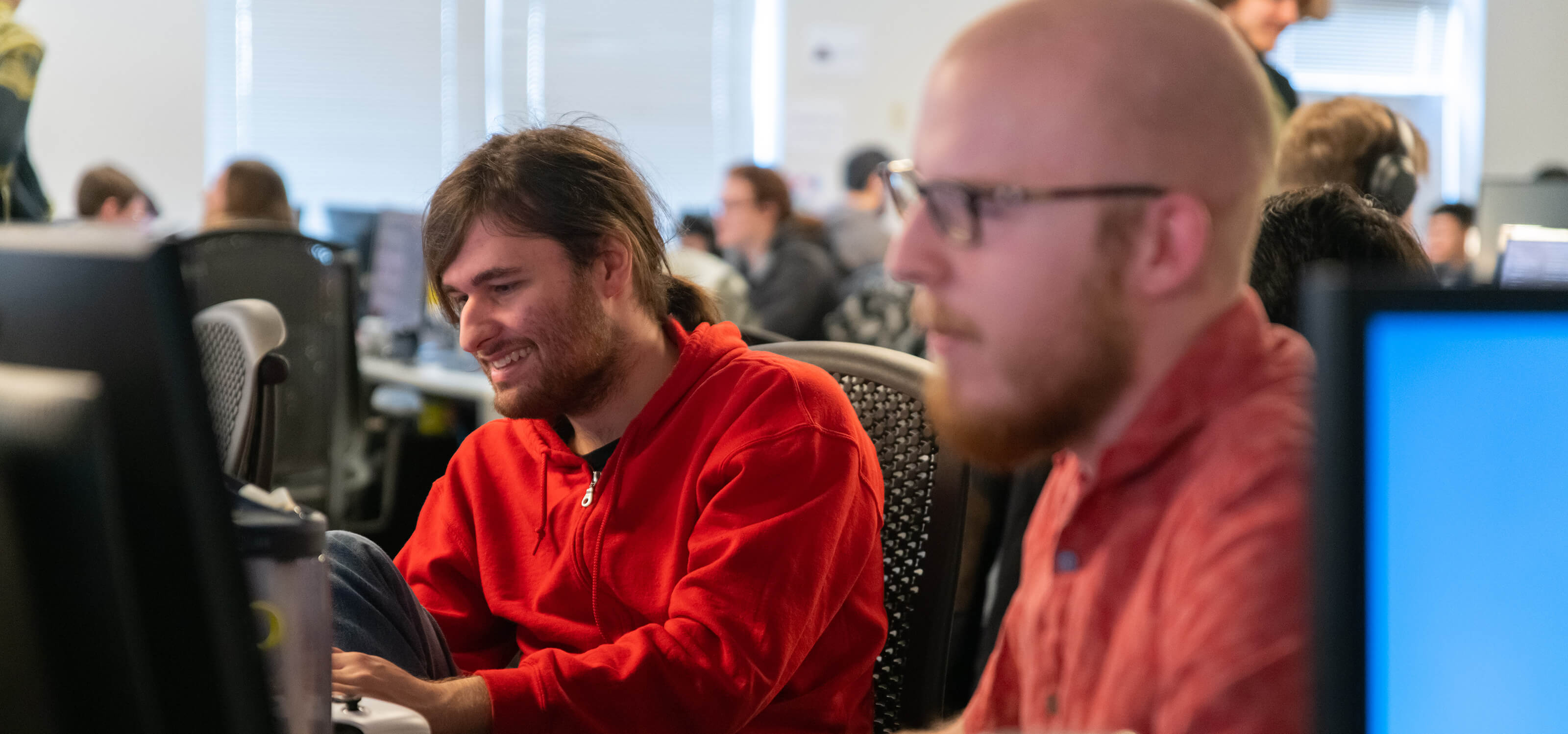 A DigiPen student in a red hoodie smiles as he works on his game project in the Tesla production lab.