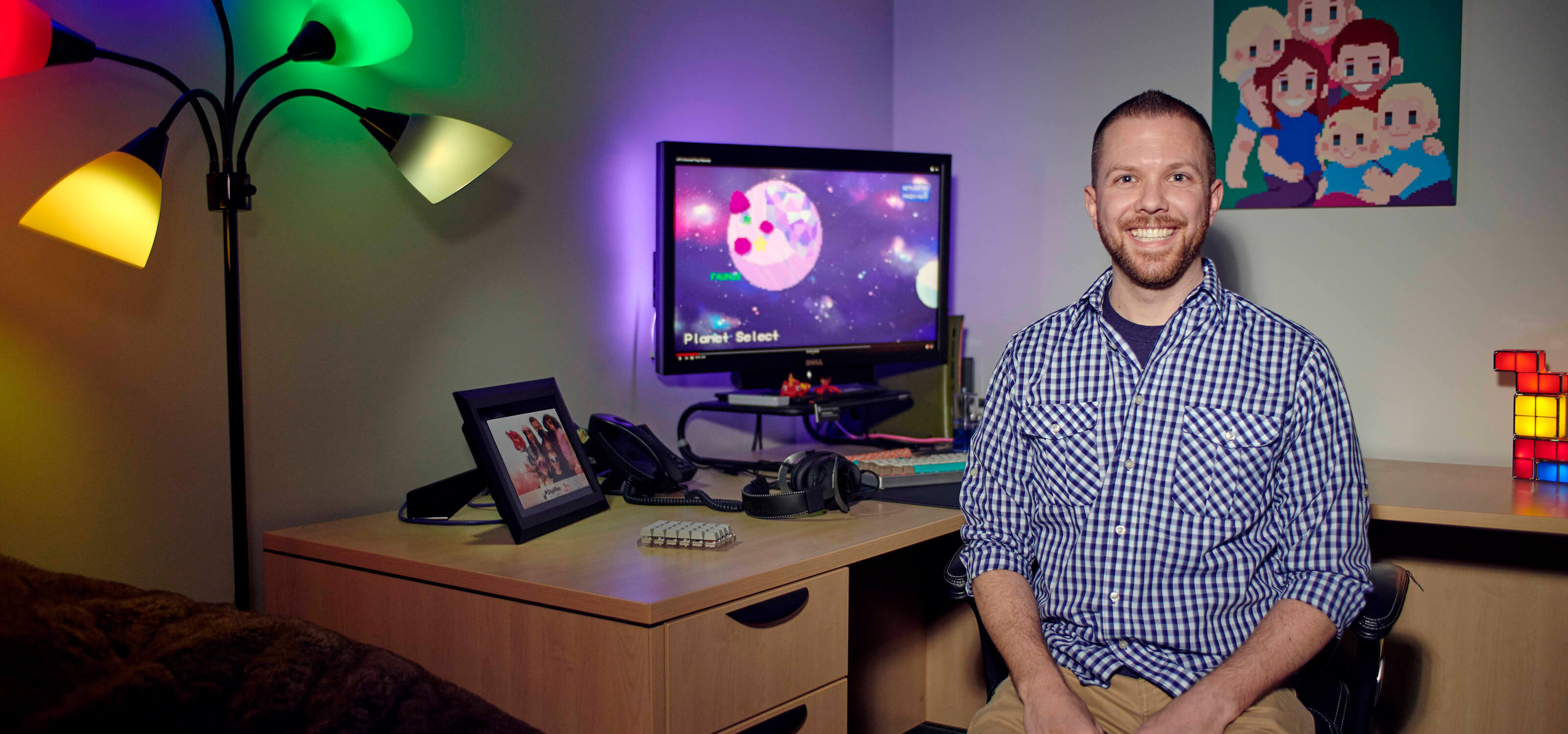 Justin Chambers smiles facing camera at his office desk, dark room decorated with objects and illuminated by colorful lamps.